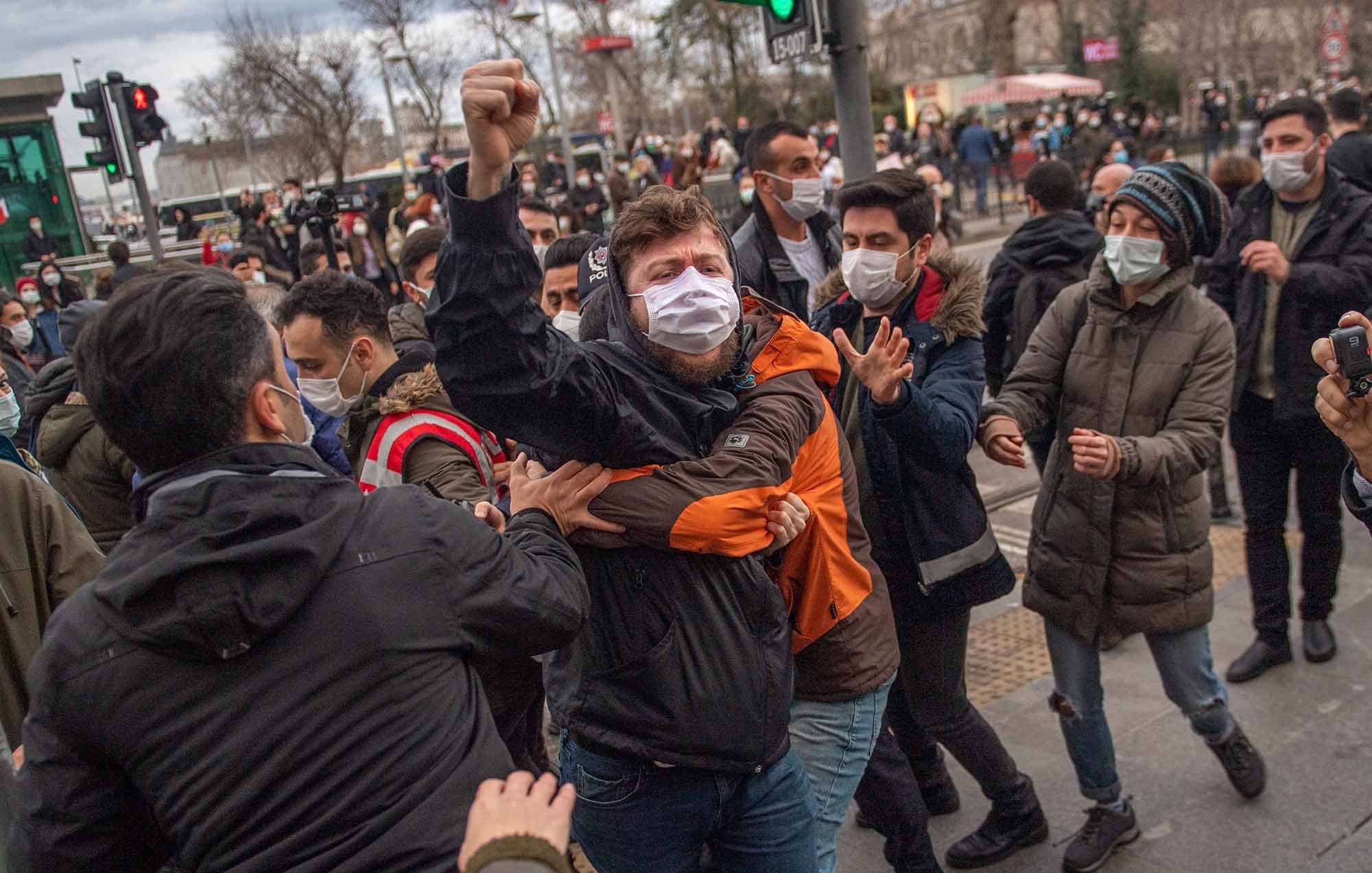 تظهر الصورة مجموعة من الأشخاص في مشهد حيوي، حيث يبدو أن هناك احتجاجًا أو تجمعًا. بعضهم يرتدي الكمامات، مما يشير إلى الالتزام بإجراءات السلامة. في المقدمة، يُلاحظ شاب يرفع يده في إشارة تعبيرية، بينما يتداخل الآخرون حوله، مما يعكس حالة من التوتر أو الحماس. الخلفية تضم تفاصيل حضرية، مثل إشارات المرور والمباني، مما يوحي بأن المكان قد يكون في مدينة كبيرة.