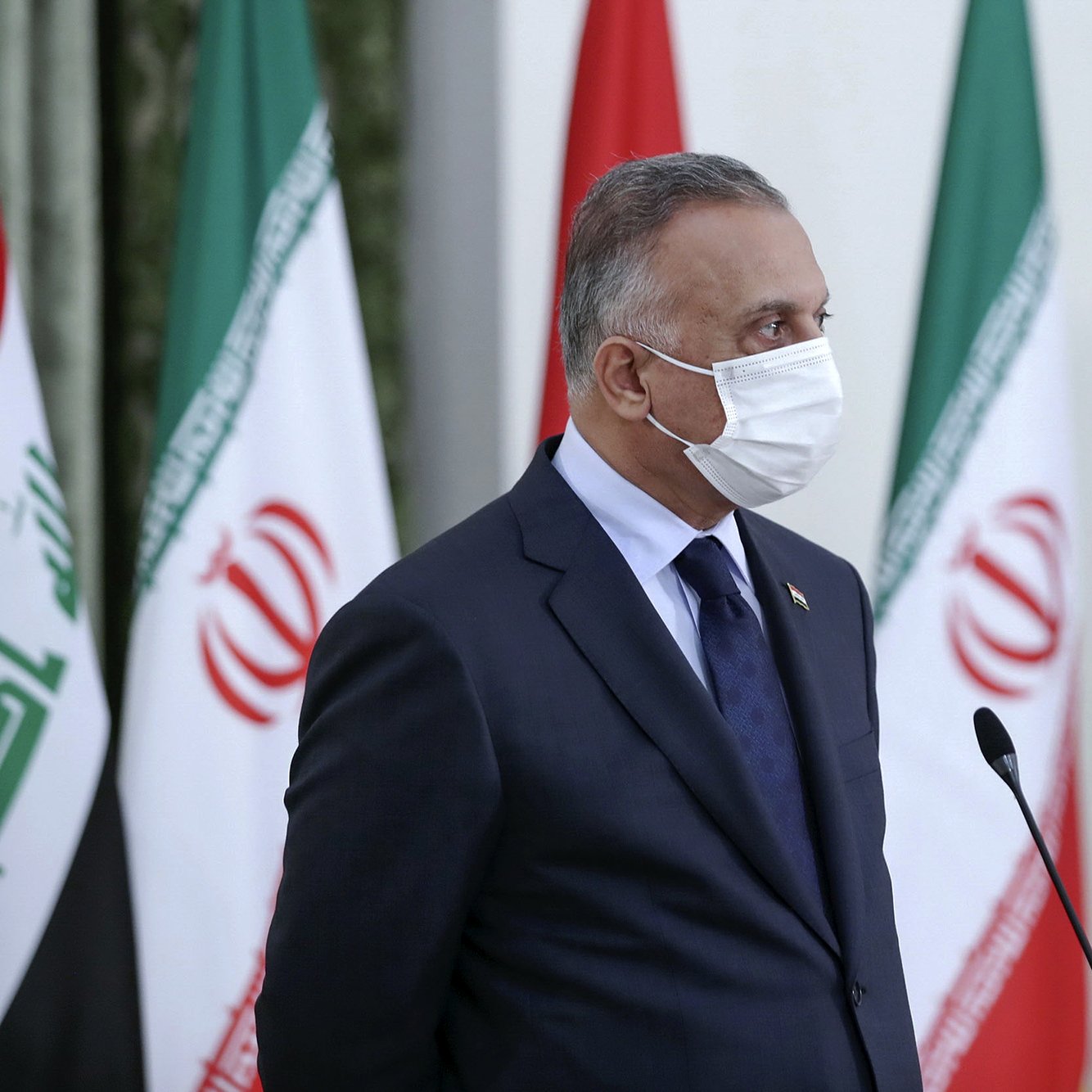 در این تصویر، مردی با روپوش رسمی و ماسک صورت دیده می‌شود. او در حال ایستاده و به یک سمت نگاه می‌کند. پشت او پرچم‌های ایران و عراق در ردیف قرار گرفته‌اند. فضا به نظر رسمی و جدی است، که نشان‌دهنده یک رویداد یا کنفرانس است.