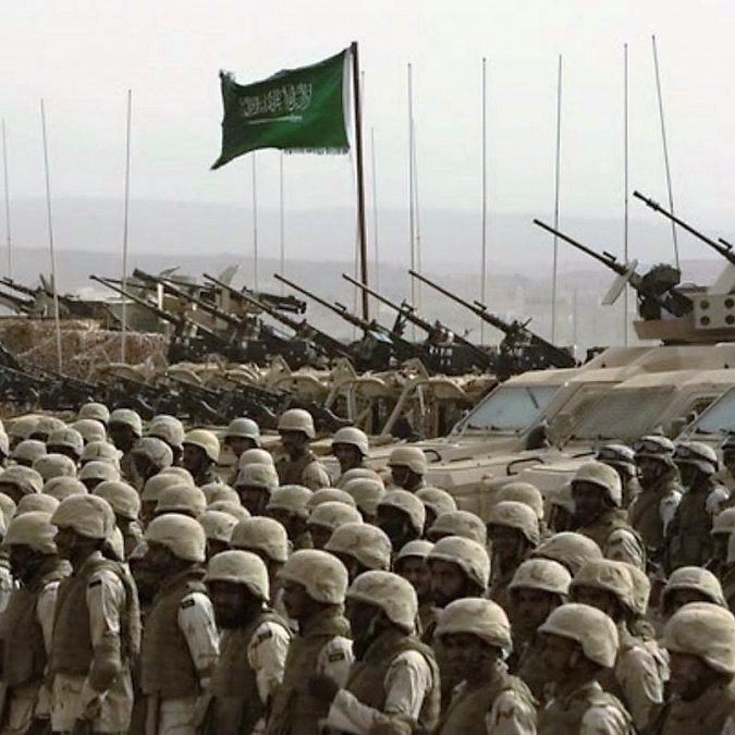 L'image montre un groupe de soldats en uniforme, alignés et mobilisés. En arrière-plan, on aperçoit des véhicules militaires et des tanks, ainsi que plusieurs canons pointés vers l'avant. Une grande bannière verte, symbolisant l'Arabie Saoudite, flotte au sommet. Le paysage est désertique, avec une ambiance d'entraînement ou de déploiement militaire.