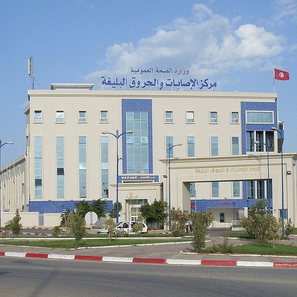 L'image montre un bâtiment moderne, probablement un centre de santé ou une institution gouvernementale. La façade est de couleur claire avec des éléments architecturaux en bleu. On y voit des fenêtres de différentes tailles et une grande enseigne en arabe et en français. Un drapeau tunisien flotte sur le toit, ce qui indique que le bâtiment est situé en Tunisie. L'environnement semble soigné, avec un espace urbain bien aménagé autour du bâtiment.