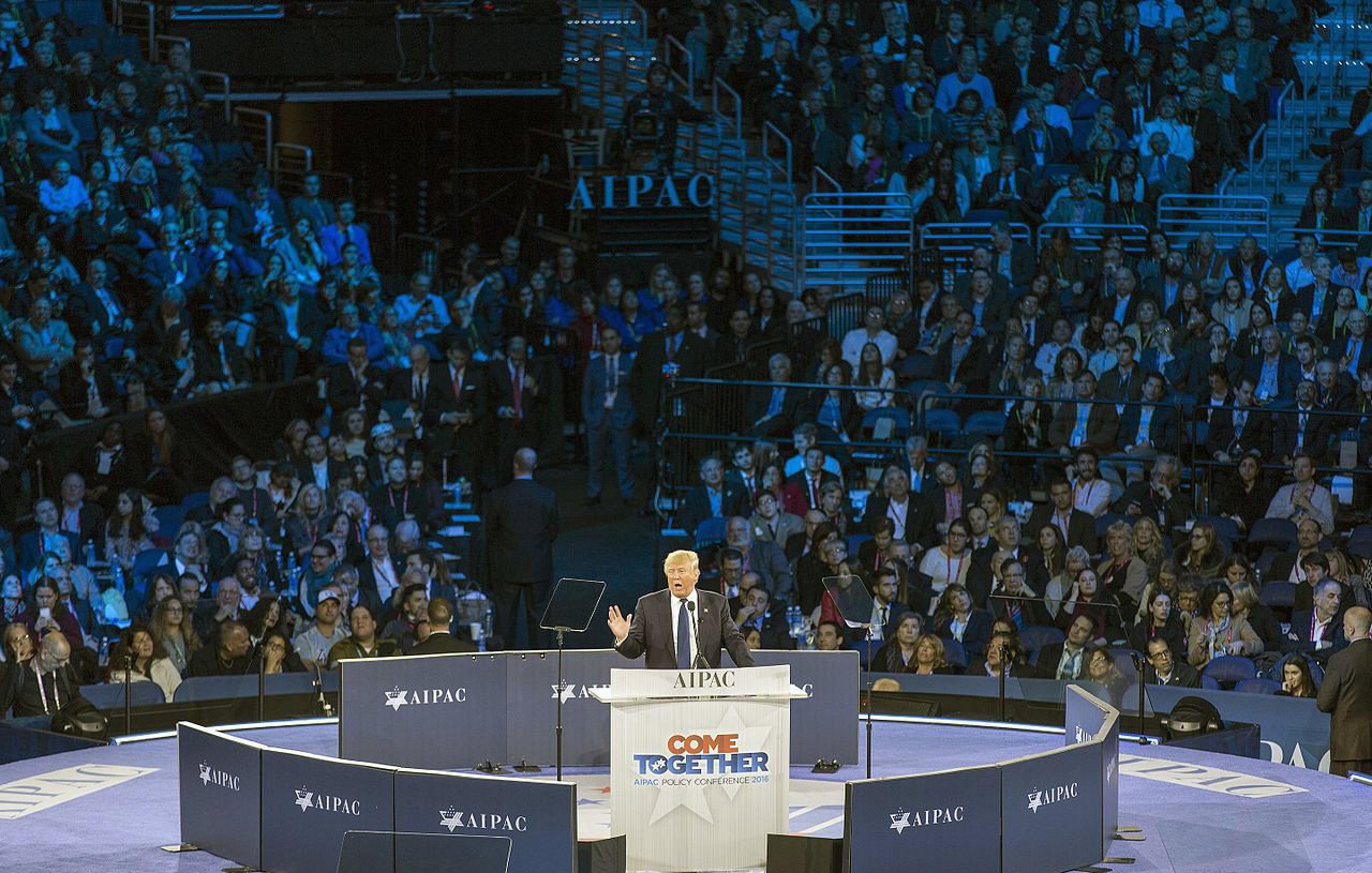 تظهر الصورة مشهداً لمؤتمر معبد مخصص لجمعية "AIPAC"، حيث يتحدث متحدث على المنصة أمام جمهور كبير. المنصة محاطة بأعلام وشعارات الجمعية، كما أن الإضاءة تلقي ظلالاً زرقاء على الحضور. يبدو أن المتحدث يناقش قضية مهمة، حيث يبدو الحضور متفاعلاً مع حديثه.