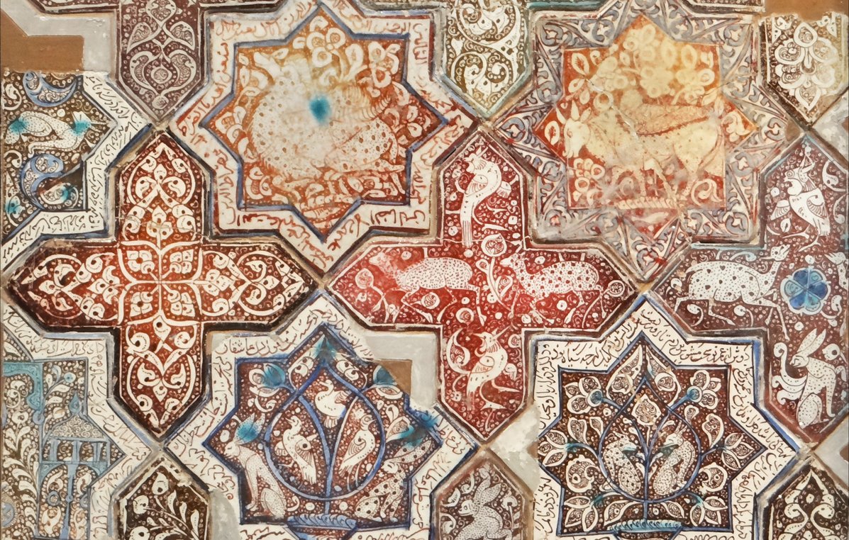 L'image présente un motif complexe et orné, typique de l'art islamique. On y voit des formes géométriques et des étoiles, réalisées dans des teintes richement variées allant des rouges aux bruns, avec des touches de bleu et de blanc. Les motifs sont décorés de détails floraux et d'animaux stylisés, ajoutant à la richesse visuelle de l'ensemble. Cette composition évoque une harmonie artistique ainsi qu'un savoir-faire traditionnel, caractéristique des mosaïques ou des carreaux décoratifs.