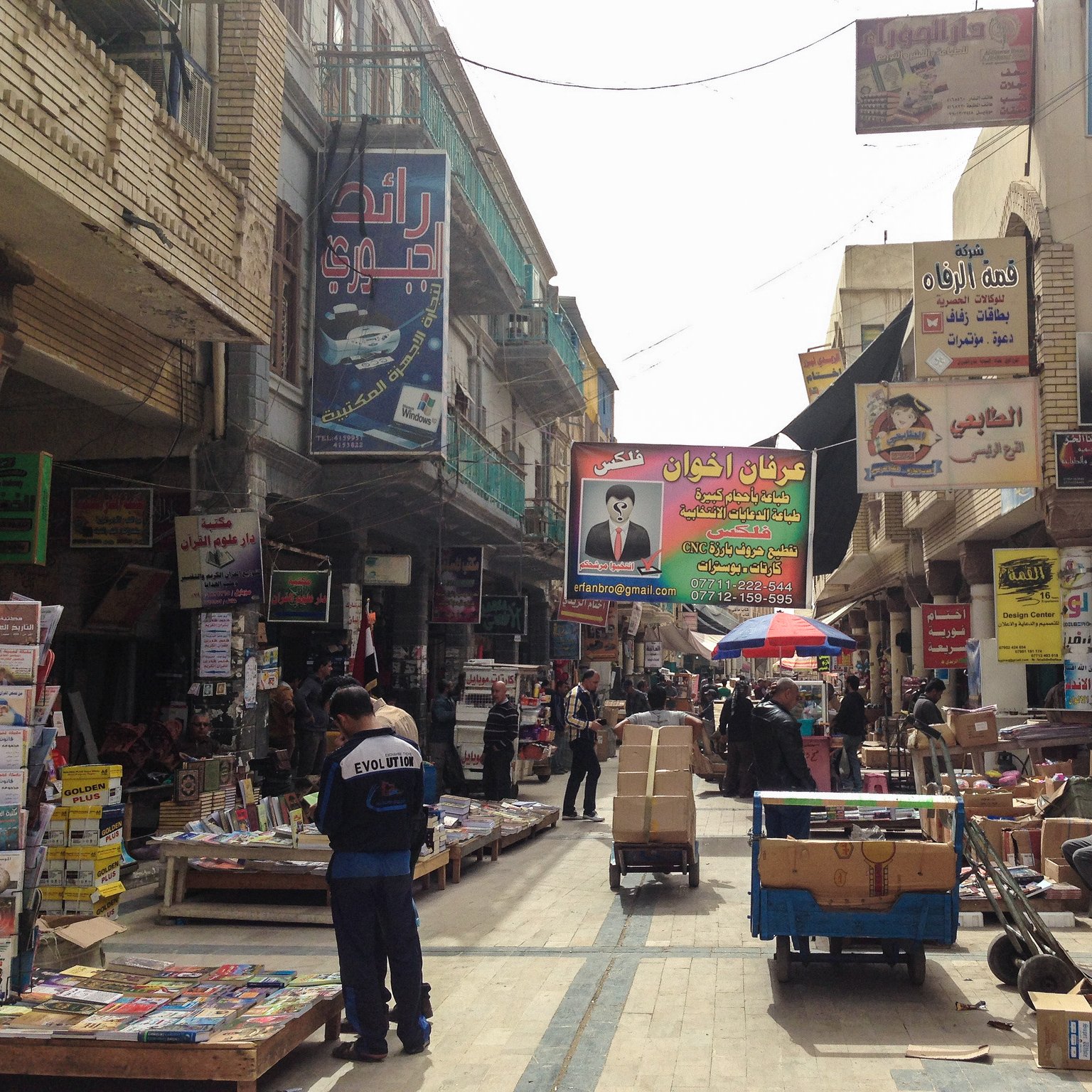 L'image montre une rue animée, typique d'un marché urbain dans une ville. On peut voir des étals et des magasins de part et d'autre, présentant divers produits, avec des affiches et des panneaux publicitaires colorés accrochés aux murs. Certaines personnes sont visibles, allant et venant, tandis que d'autres semblent être en train de travailler, probablement en déchargeant des marchandises. Le sol est souvent encombré de caisses et d'autres objets, créant une ambiance vivante et dynamique. Des parasols sont également présents, offrant un peu d'ombre dans cette rue fréquentée.