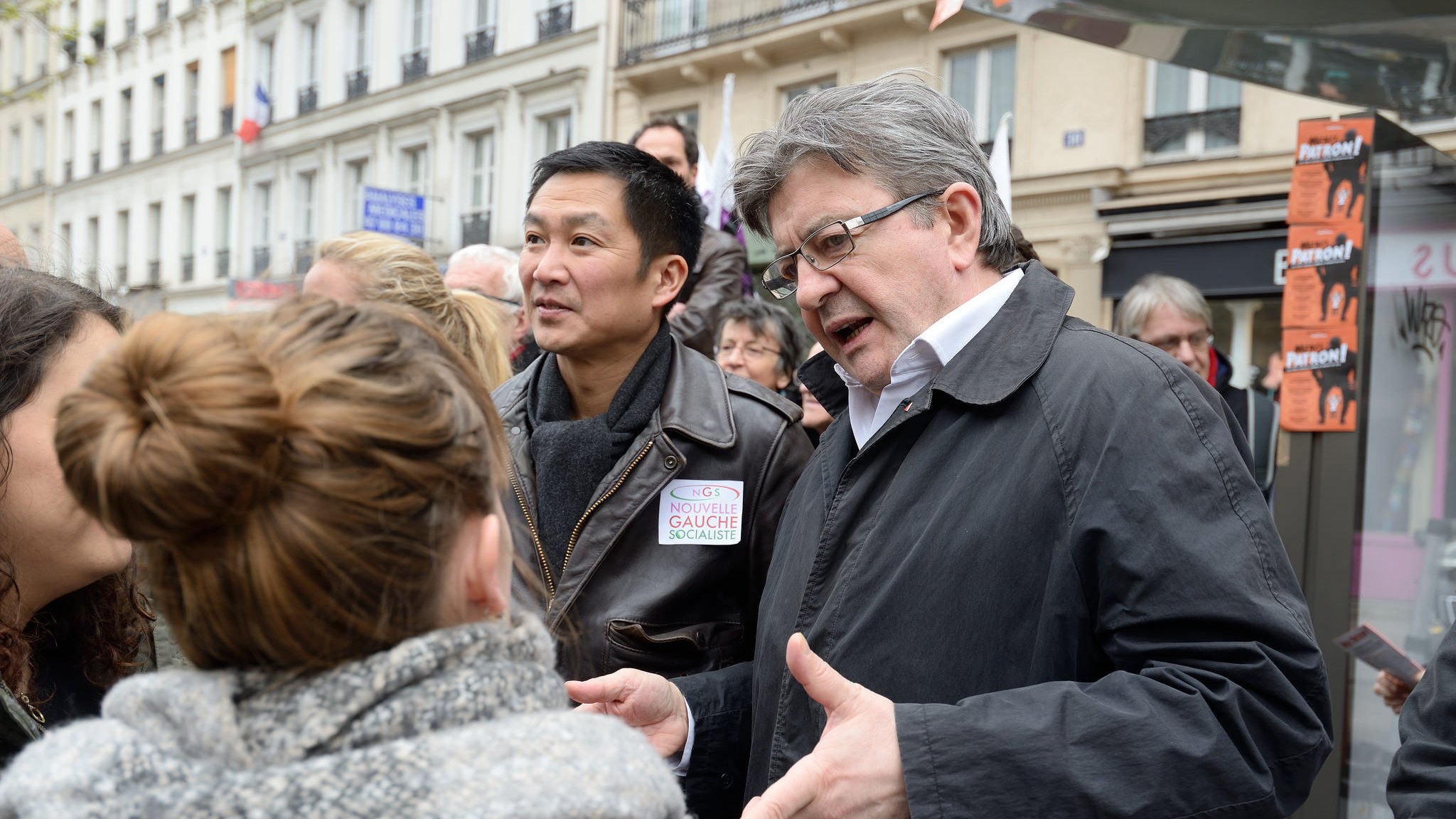 L'image montre un groupe de personnes en train de discuter dans une rue, probablement lors d'un événement public. On peut voir deux hommes au premier plan, l'un d'eux portant une veste sombre. Ils semblent engagés dans une conversation avec des femmes. L'ambiance semble dynamique et interactive, suggérant un échange d'idées ou de points de vue. Les bâtiments en arrière-plan indiquent qu'ils se trouvent dans un environnement urbain.