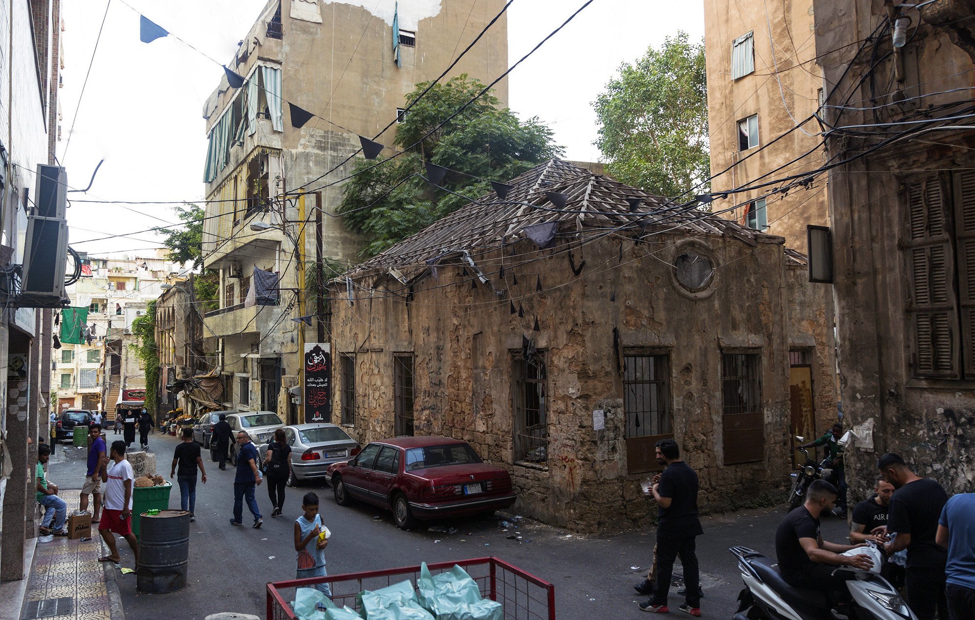 L'image montre une ruelle étroite dans une ville, probablement au Liban. On peut y voir des bâtiments anciens avec des murs en pierre usée par le temps. Certains bâtiments semblent en mauvais état, tandis que d'autres sont légèrement rénovés. La rue est animée avec des personnes qui marchent, certaines d'entre elles se tenant près de véhicules stationnés. Des banderoles pendant au-dessus de la rue ajoutent une touche de couleur. En général, l'atmosphère semble à la fois vivante et marquée par l'histoire.