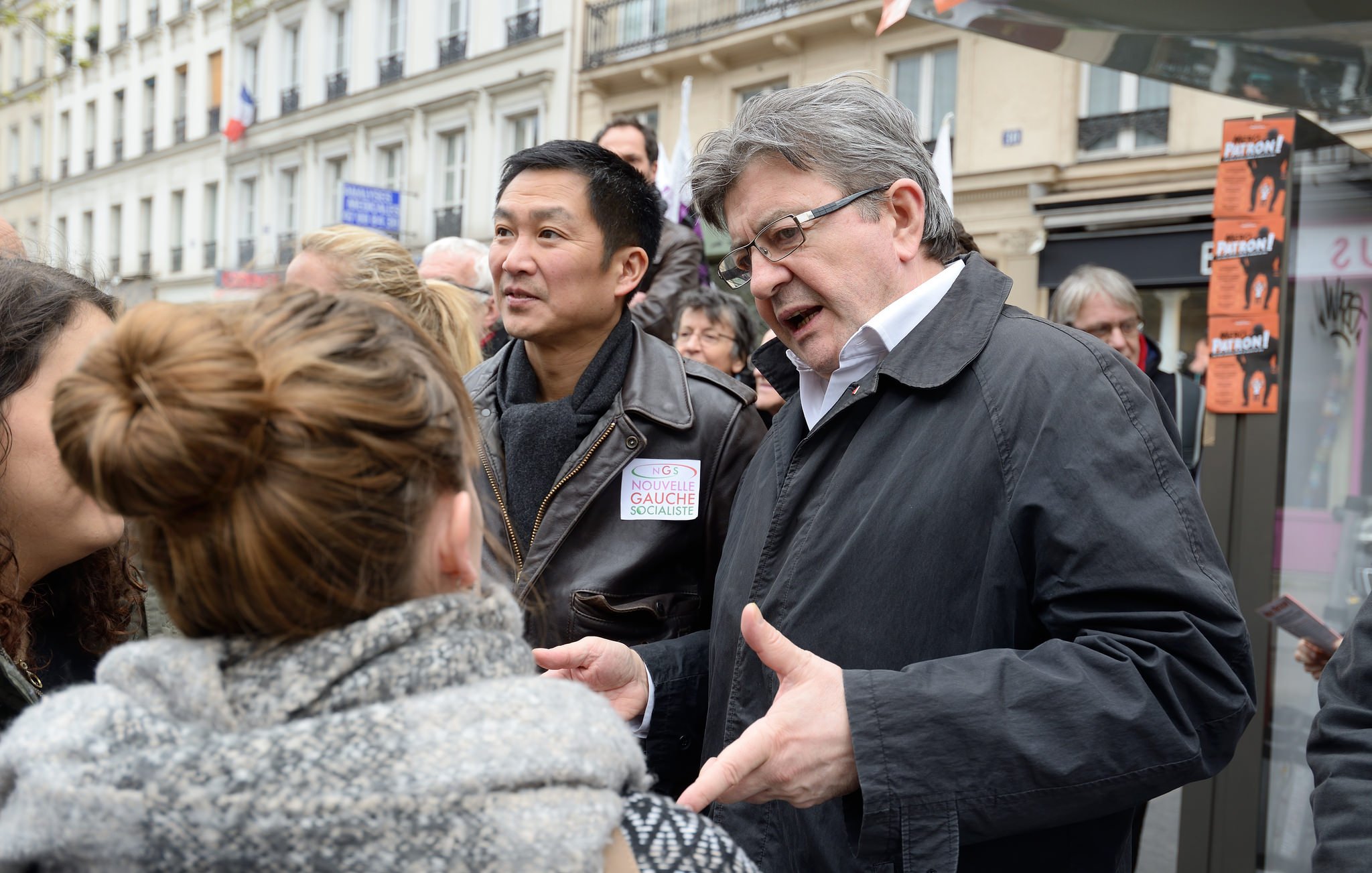 L'image montre un groupe de personnes en train de discuter dans une rue, probablement lors d'un événement public. On peut voir deux hommes au premier plan, l'un d'eux portant une veste sombre. Ils semblent engagés dans une conversation avec des femmes. L'ambiance semble dynamique et interactive, suggérant un échange d'idées ou de points de vue. Les bâtiments en arrière-plan indiquent qu'ils se trouvent dans un environnement urbain.