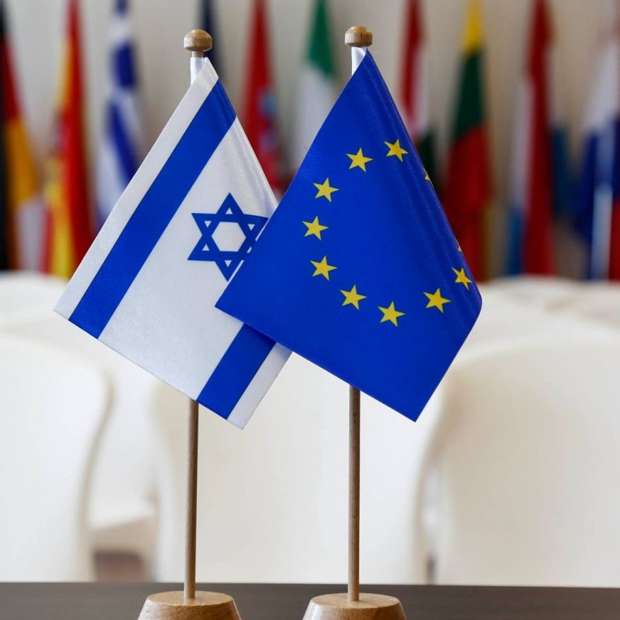 L'image montre deux drapeaux placés sur des supports. À gauche se trouve le drapeau d'Israël, qui présente une étoile de David et deux bandes bleues. À droite se trouve le drapeau de l'Union européenne, emblématique avec des étoiles dorées disposées en cercle sur un fond bleu. En arrière-plan, on aperçoit un rassemblement de drapeaux de différents pays, suggérant un contexte international ou diplomatique. Les chaises blanches sont disposées derrière, évoquant une salle de réunion ou une conférence.