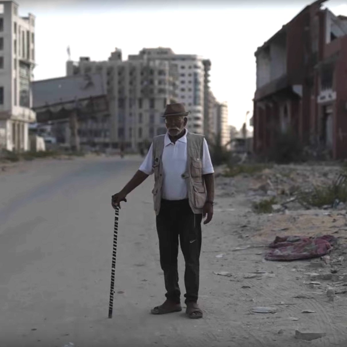Un homme âgé avec une canne se tient sur une rue déserte, entouré de bâtiments en ruine.