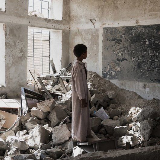 L'image montre un enfant se tenant dans une salle de classe en ruines. Le sol est jonché de débris, tandis que des morceaux de murs effondrés et des meubles renversés témoignent de la destruction. Un tableau noir, partiellement visible, porte des inscriptions, bien que l'environnement soit dégradé. La lumière pénètre à travers les fenêtres, créant un contraste avec l'atmosphère sombre et désolée de la pièce. Cette scène évoque une profonde sensation de perte et de désespoir.