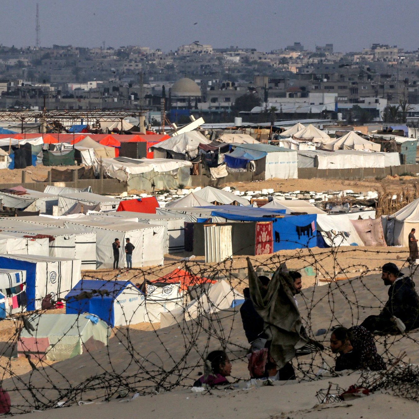 L'image montre un camp de réfugiés avec de nombreuses tentes de différentes couleurs, principalement blanches et bleues, s'étendant sur un terrain désertique. À l'arrière-plan, on aperçoit une ville avec des bâtiments. On peut également distinguer des personnes assises, ainsi que des barbelés entourant le camp, ce qui souligne une ambiance de sécurité précaire et d'incertitude. L'ensemble évoque une situation difficile et des conditions de vie sommaires.