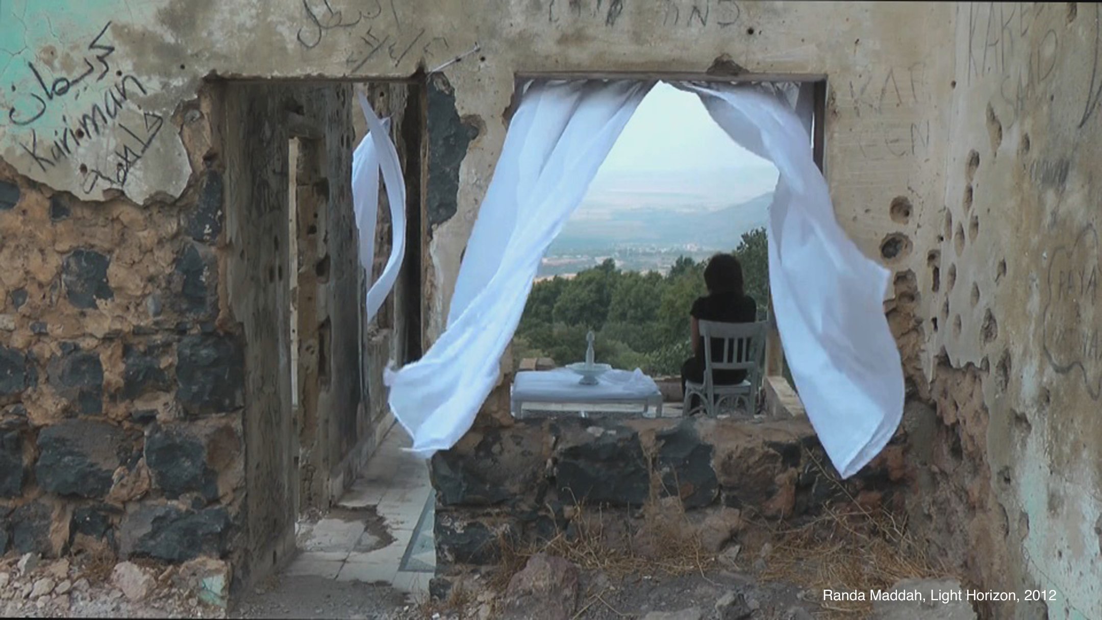 L'image montre une scène à l'intérieur d'un bâtiment partiellement ruiné. On peut voir une fenêtre encadrée par des rideaux blancs flottants. À travers cette fenêtre, un paysage verdoyant s'étend avec des collines au loin. À l'intérieur, une chaise blanche est placée près d'une petite table, créant une atmosphère de calme et de solitude. Les murs sont marqués par des graffitis, ajoutant une dimension de mémoire et d'histoire à l'espace.