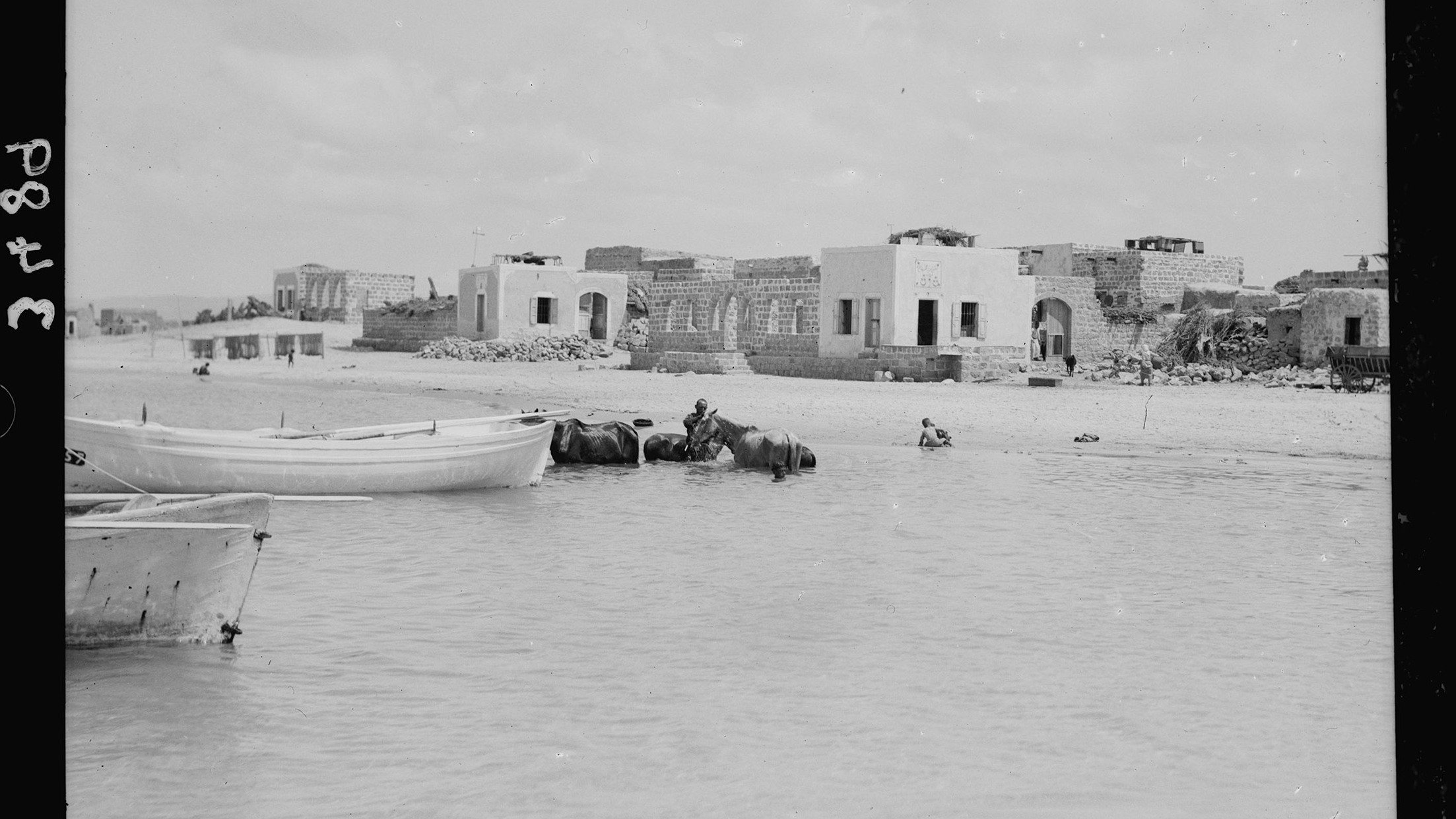 تظهر في الصورة مشهداً ساحلياً يضم مجموعة من المراكب الصغيرة الراسية على المياه. في الخلفية، توجد مبانٍ قديمة إلى جانب بعض الهياكل المتبقية، ما يدل على وجود مجتمع ساحلي سابق. يُرى بعض الأشخاص، بما في ذلك شخص يركب على ظهر حيوان، مما يعطي انطباعًا عن الحياة اليومية في هذا المكان. تتسم السماء بالغيوم المتفرقة، مما يوحي بأن الصورة ربما تم التقاطها في يوم مشمس.