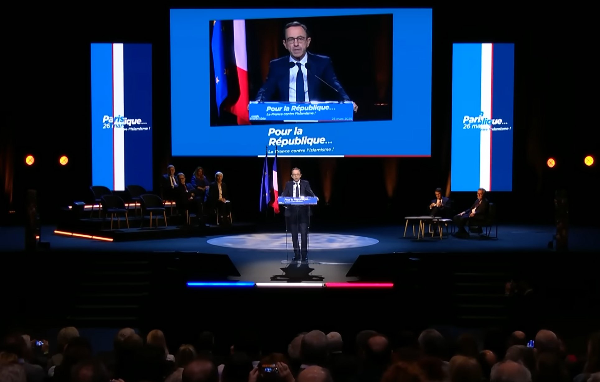 Un homme parle sur scène, avec des drapeaux français en arrière-plan. 