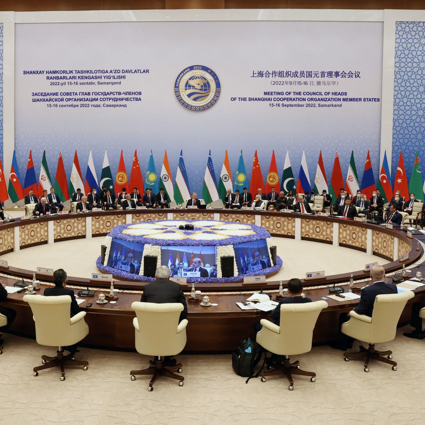 The image depicts a large conference room set up for a high-level meeting, with a circular table at the center surrounded by delegates. Various flags representing different countries are displayed around the room, indicating a multinational gathering. Attendees, likely officials or representatives, are seated and engaged in discussion. The backdrop features stylized graphics and text, suggesting it is related to a significant international conference, possibly focusing on cooperation among member states. The atmosphere appears formal and collaborative.