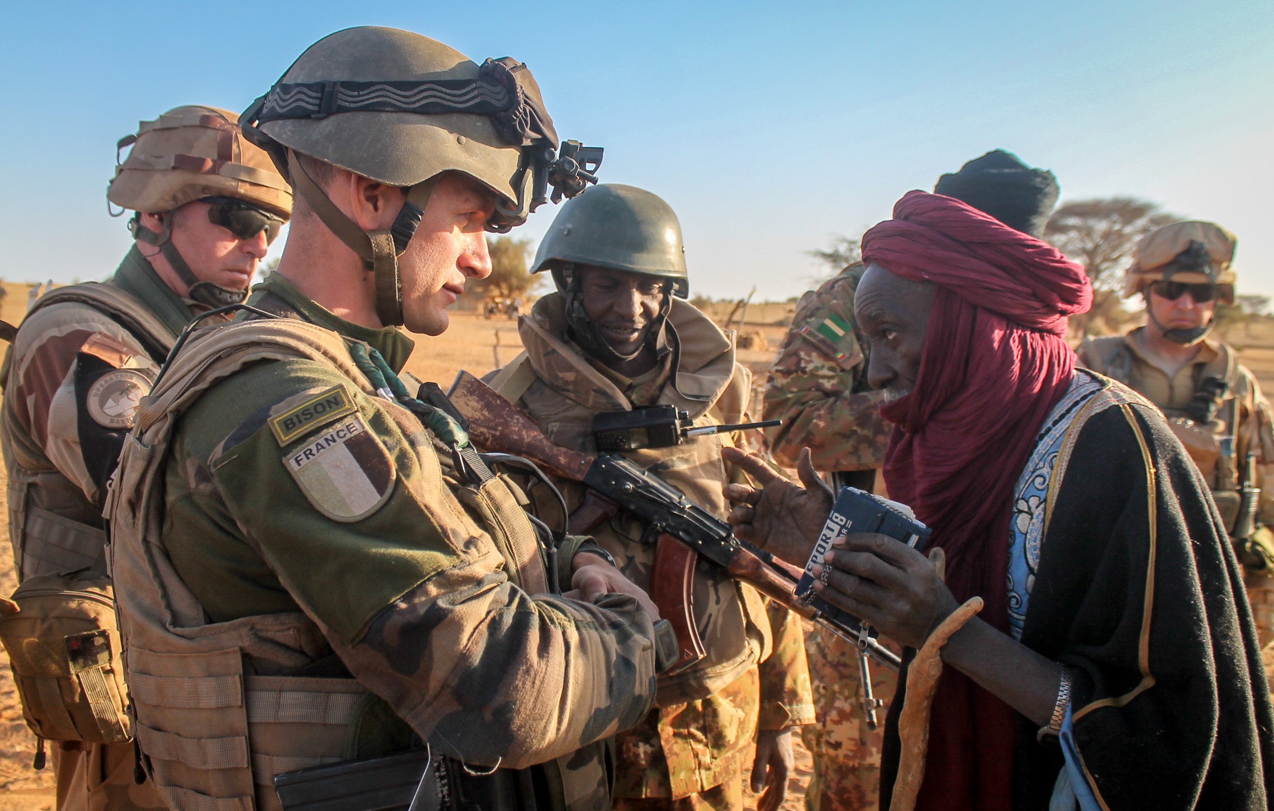 L'image montre un soldat en uniforme militaire, probablement français, interagissant avec un homme âgé, qui semble local. Le soldat est armé et porte un gilet pare-balles ainsi qu'un casque, tandis que l'homme a des vêtements traditionnels. En arrière-plan, d'autres militaires, également en uniforme, observent la scène. Le cadre semble désertique, typique d'une région saharienne, avec un ciel dégagé et un sol sablonneux. L'interaction semble empreinte de respect et d'échange culturel.