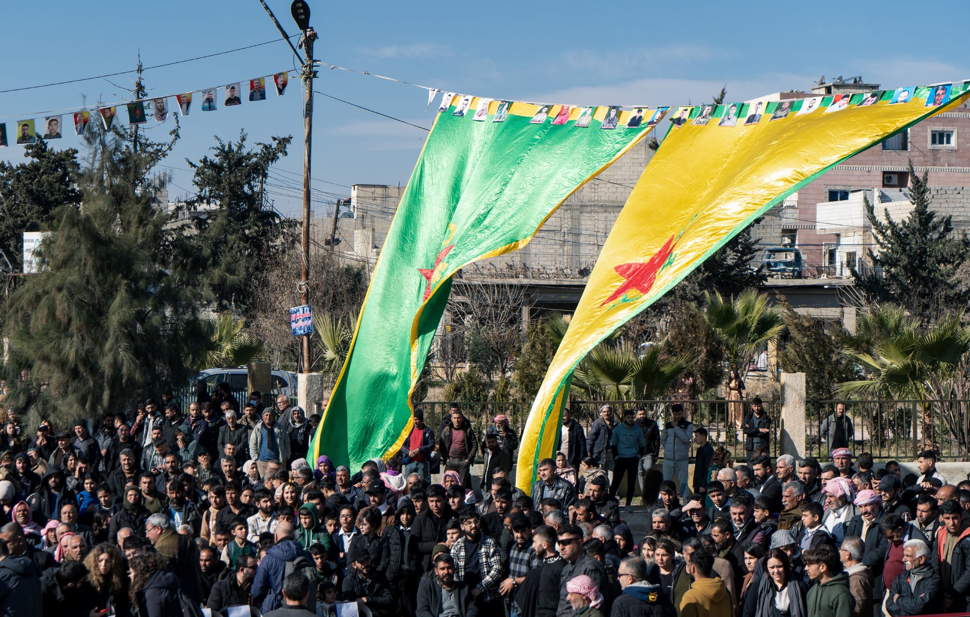 L'image montre une foule rassemblée sous de grandes drapeaux verts et jaunes, portant une étoile rouge. En arrière-plan, des bâtiments et des arbres sont visibles. Des banderoles sont accrochées au-dessus des personnes, indiquant une célébration ou un rassemblement communautaire. L'atmosphère semble vibrante et engagée, avec un grand nombre de participants.