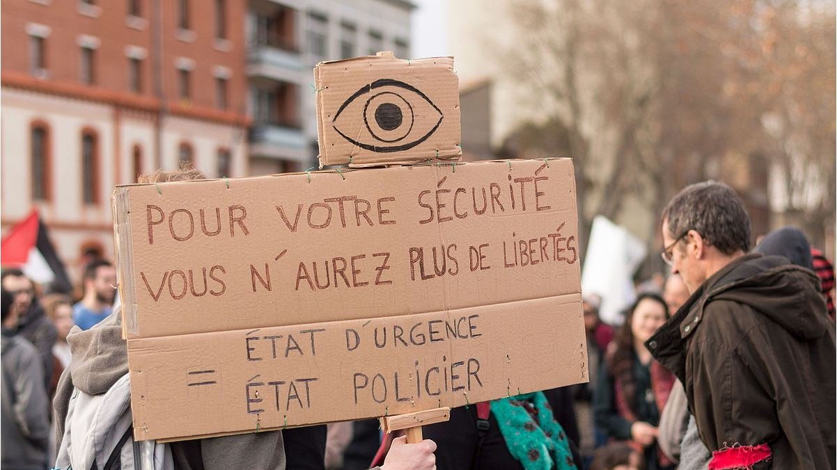 L'image représente une manifestation où une personne tient une pancarte en carton. Sur cette pancarte, on peut lire un message dénonçant la restriction des libertés au nom de la sécurité. Le symbole d'un œil est visible en haut de la pancarte, renforçant le thème de la surveillance. Les participants de la manifestation semblent mobilisés, et il y a une ambiance de protestation dans l'air.