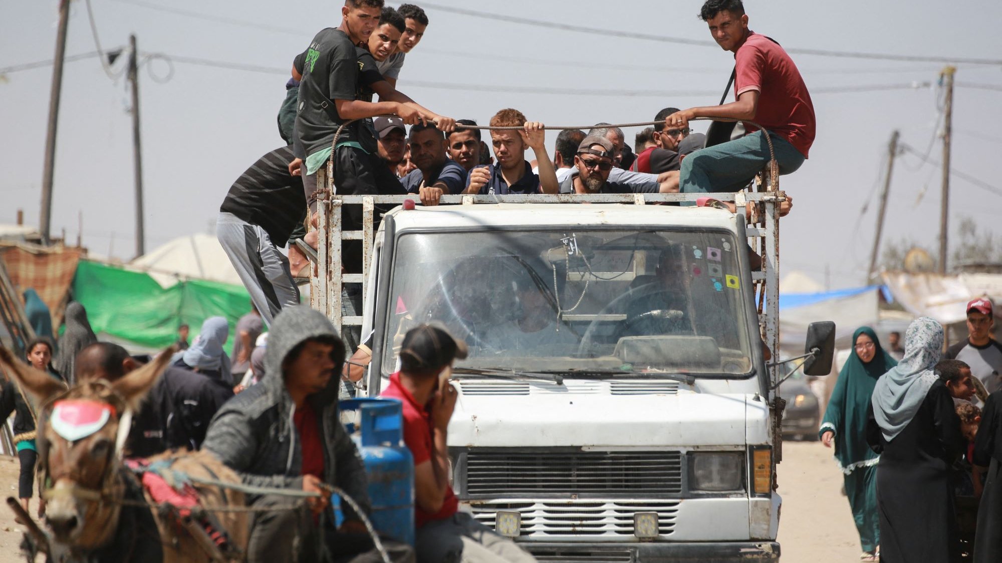 L'image montre un groupe de personnes assises sur le toit d'un camion, tandis que d'autres se trouvent à proximité, y compris des personnes à pied et une charrette tirée par un cheval. L'environnement semble être un endroit animé, avec des tentes et des structures visibles en arrière-plan. On peut voir des fils électriques et des montagnes de déchets, ce qui suggère une atmosphère informelle ou de camp. La scène dégage une impression de communauté et de vie quotidienne dans un contexte peut-être difficile.