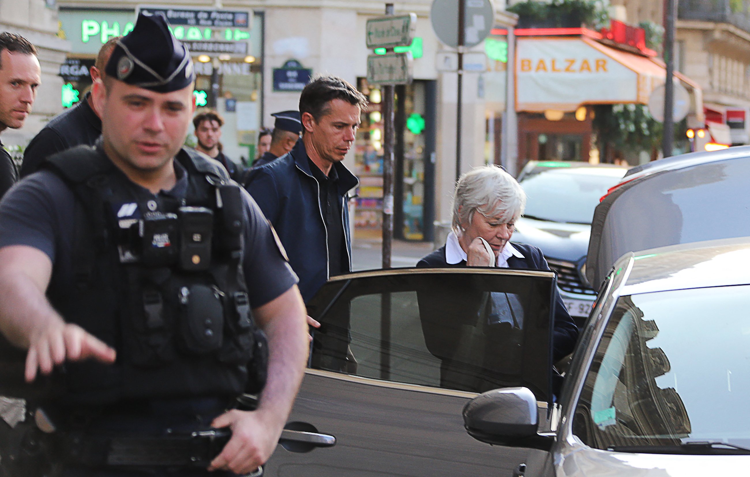 L'image montre une scène urbaine avec plusieurs personnes dans une rue. On peut voir un policier en uniforme à l'avant, surveillant la situation. À côté de lui, un homme et une femme semblent sortir d'une voiture. La femme est habillée de manière formelle et semble pensive. En arrière-plan, il y a plusieurs enseignes de magasins qui ajoutent à l'ambiance de la ville. Les lumières et l'architecture confèrent une atmosphère animée à la scène.