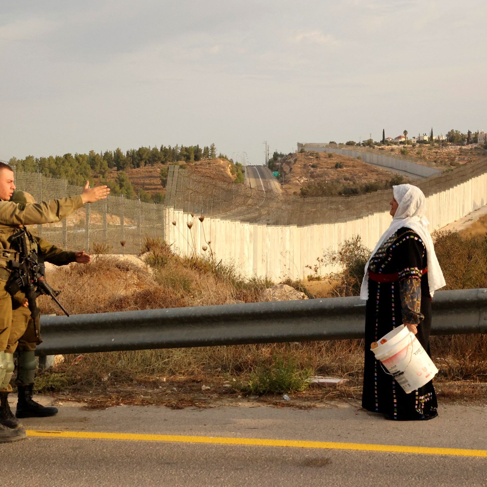 L'image montre un soldat en uniforme qui semble communiquer avec une femme vêtue de manière traditionnelle, tenant un seau. En arrière-plan, on aperçoit un mur ou une barrière qui sépare les deux côtés, avec des collines et des bâtiments au loin. L'environnement semble rural, avec de la végétation et une route. Cette scène évoque un contexte de conflit ou de tension dans une région marquée par des divisions.