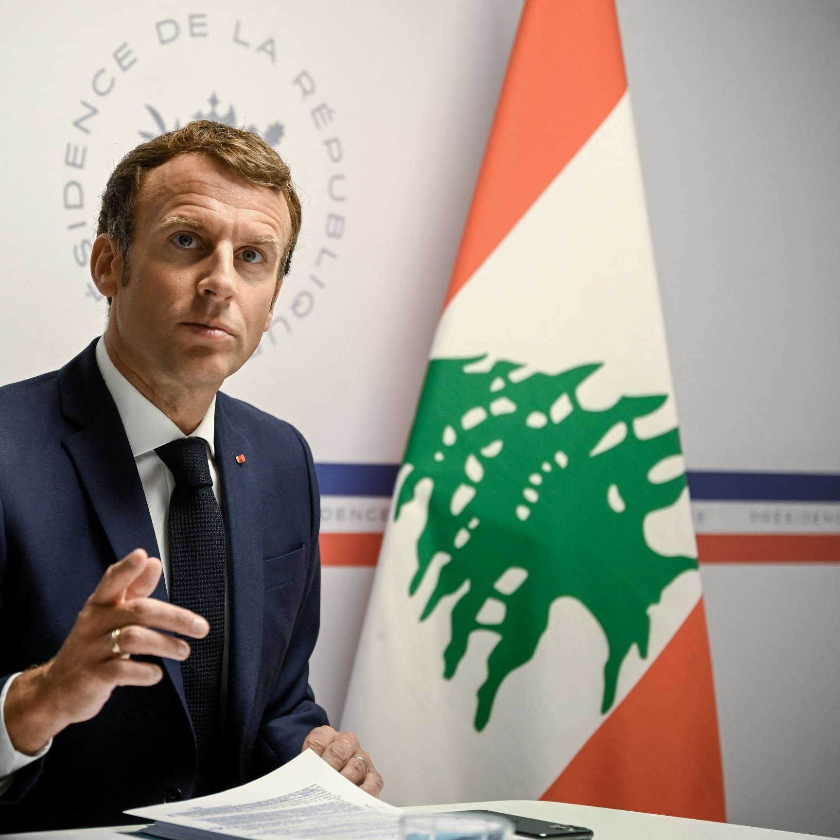 در تصویر، یک مرد با موی تیره و لباس رسمی (کت و شلوار) دیده می‌شود که به سمت دوربین نگاه می‌کند. او دارای ظاهری جدی و متفکر است و با دستش در حال بیان نقطه نظری است. در پس‌زمینه، پرچم لبنان و نشان رسمی نشان داده شده است. به نظر می‌رسد که این فرد در یک جلسه یا کنفرانس خبری مشغول سخنرانی است.