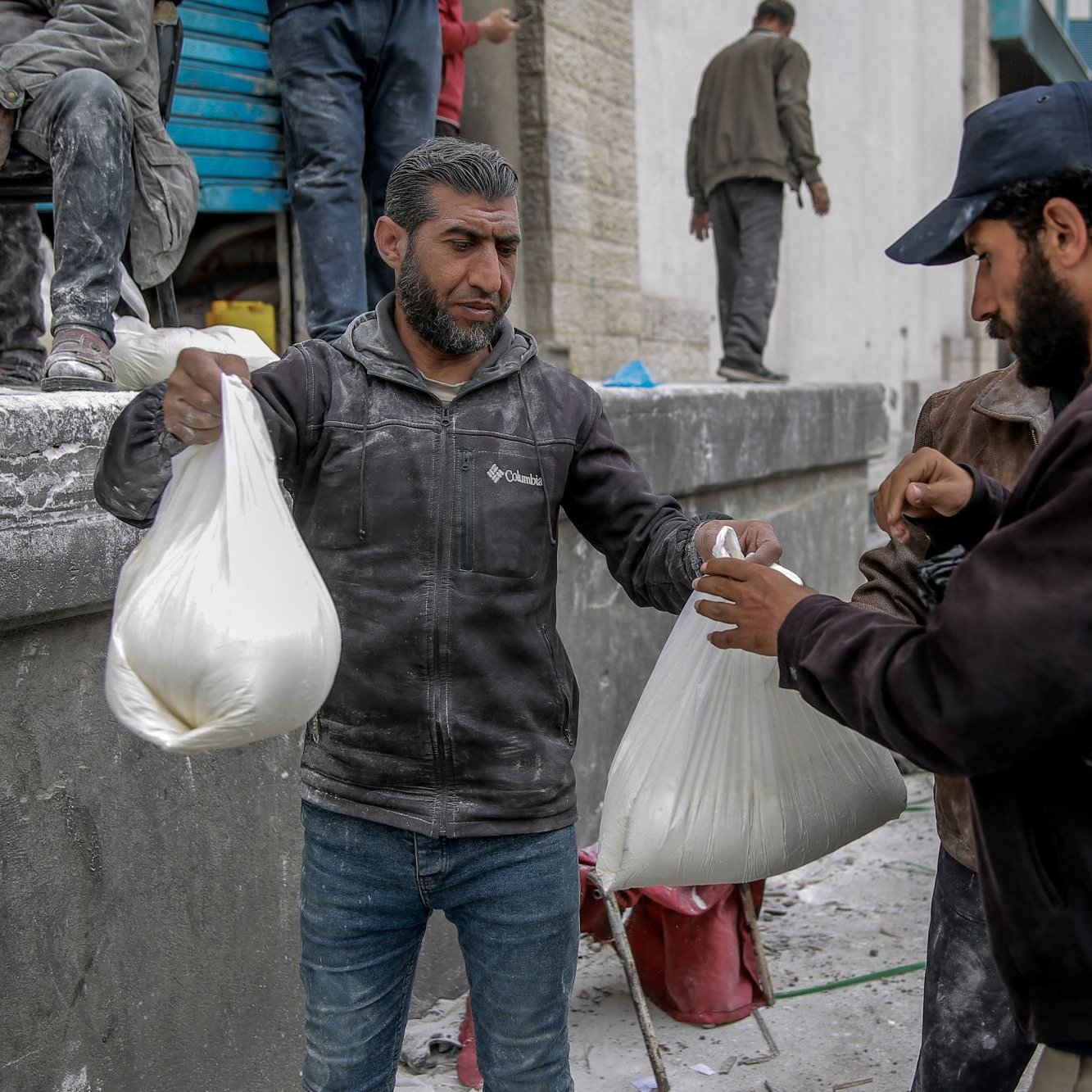 L'image montre des hommes dans un environnement urbain, apparemment en train de distribuer ou de recevoir des sacs. Ils semblent porter des vêtements de travail, certains étant couverts de poussière ou de matériau. L'ambiance suggère une activité liée à l'aide humanitaire ou à une distribution de biens, dans un contexte peut-être de reconstruction ou d'assistance. Les interactions entre les hommes semblent concentrées et sérieuses.