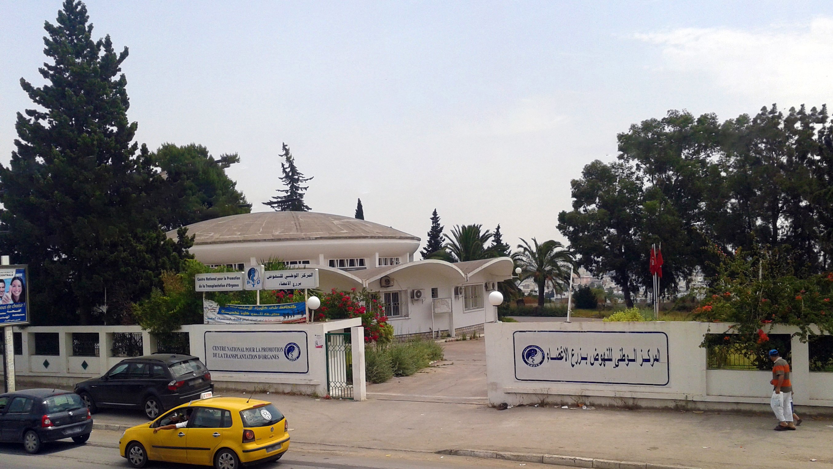 The image depicts a modern building with a dome-shaped roof, likely serving as a public institution or center. The architecture features a combination of curved and angular elements, with large windows that allow natural light inside. Surrounding the building are trees and greenery, contributing to a pleasant atmosphere. In front of the building, there are vehicles parked, including a yellow taxi, indicating it is situated near a road. There are also banners displaying text, possibly providing information about the institution. A flag can be seen on a pole, suggesting this might be a governmental or community facility. Overall, the scene conveys a sense of public service in a well-maintained environment.