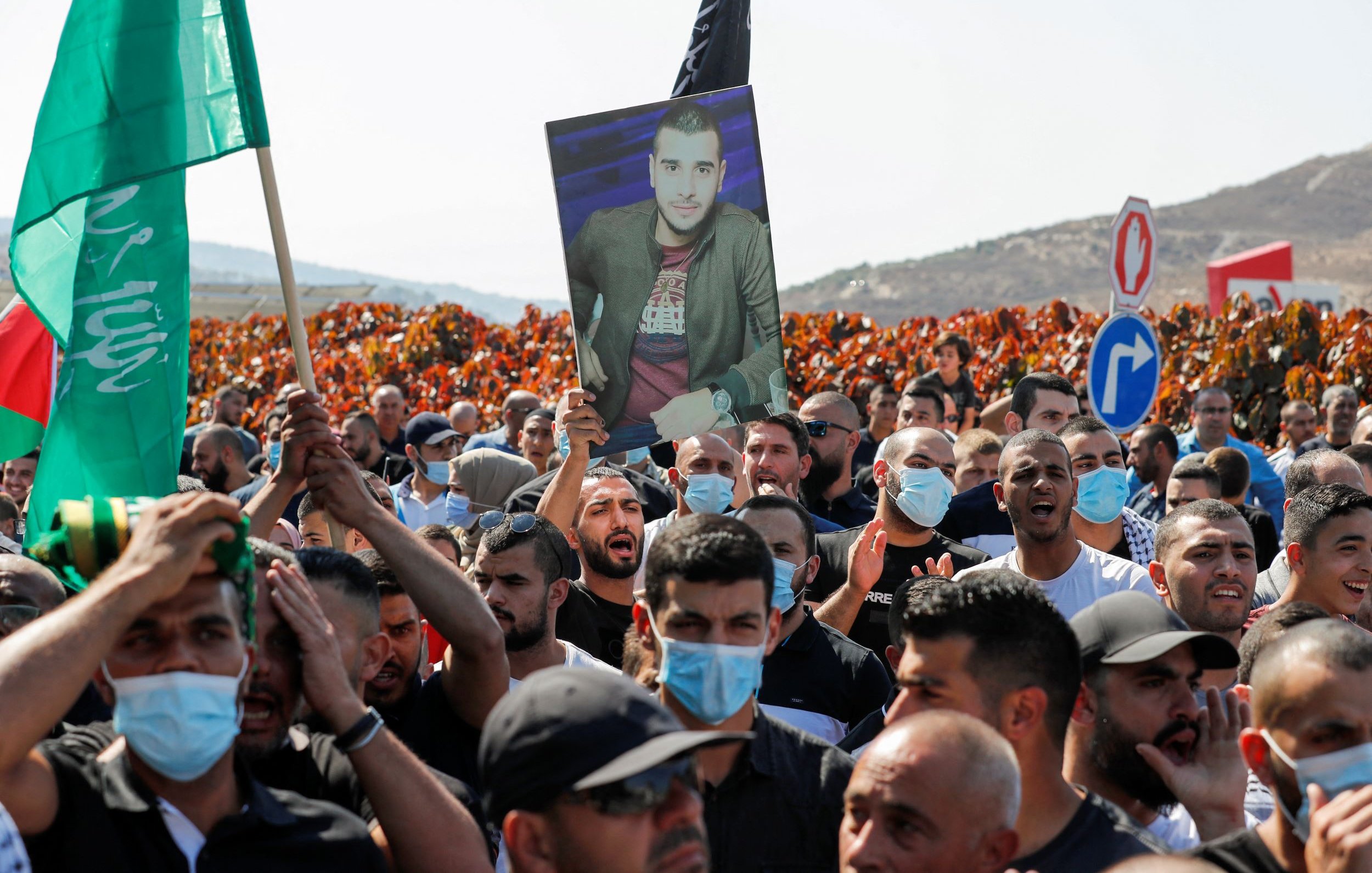 The image depicts a large crowd participating in a protest or demonstration. Many individuals are holding up flags, including green ones, and a prominent figure is holding a portrait of a man. The crowd appears to be engaged and unified, with some people wearing face masks. The background features hills, adding to the overall atmosphere of the event.