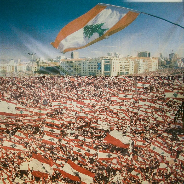 L'image montre une grande foule rassemblée, probablement lors d'une manifestation ou d'un événement public au Liban. La mer de personnes brandit de nombreux drapeaux libanais, créant un effet visuel saisissant. En arrière-plan, on aperçoit une ville moderne, ce qui suggère une ambiance vibrante. Le ciel est dégagé, ajoutant à la luminosité de la scène. Cette image évoque un sentiment de patriotisme et de solidarité parmi les participants.
