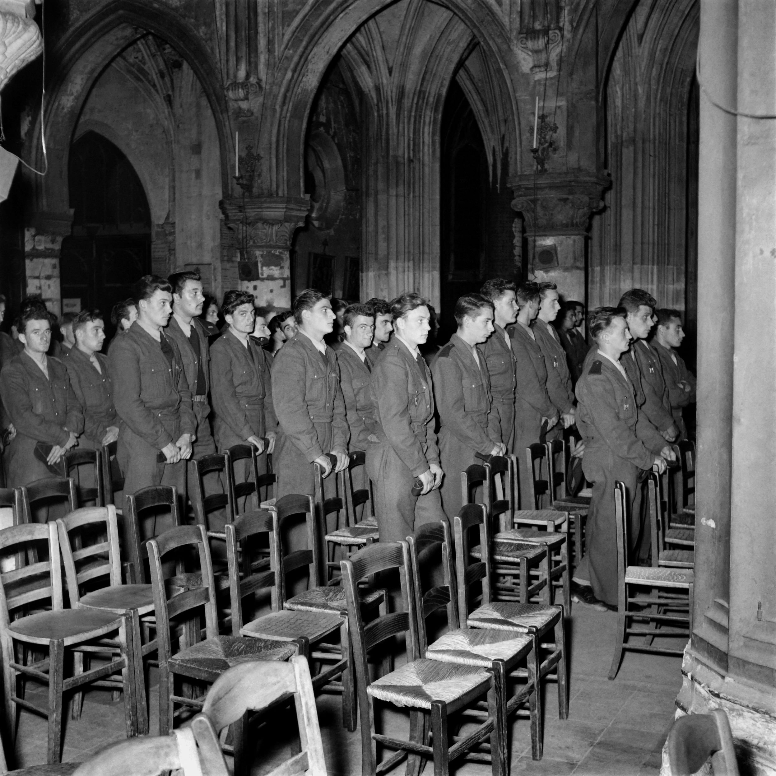 L'image montre un groupe de jeunes hommes en uniforme, probablement des soldats, se tenant debout en rang dans une église ou un bâtiment religieux. Ils semblent attentifs et concentrés, se tenant droit, dans un environnement décoré d'éléments architecturaux gothiques. Des chaises en bois sont visibles devant eux, et l'éclairage met en valeur l'atmosphère solennelle du lieu.