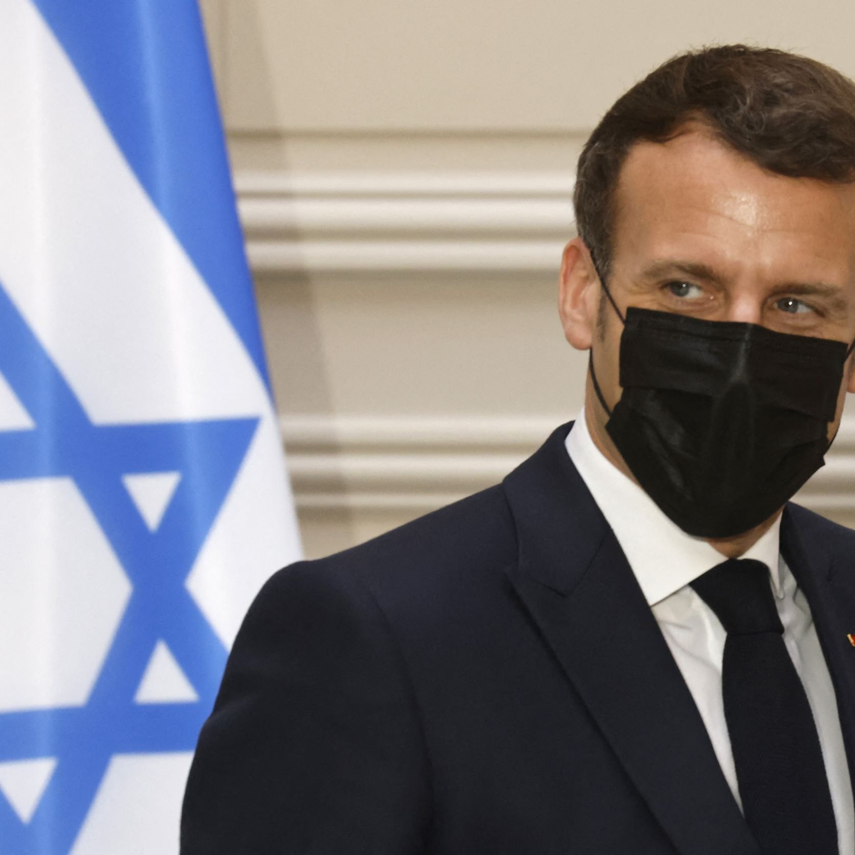 L'image montre une personne en costume, portant un masque noir, se tenant près d'un drapeau qui comporte une étoile de David. L'arrière-plan semble être un intérieur formel, peut-être un bureau ou une salle de réunion. La personne a une expression attentive et semble engagée dans une conversation.