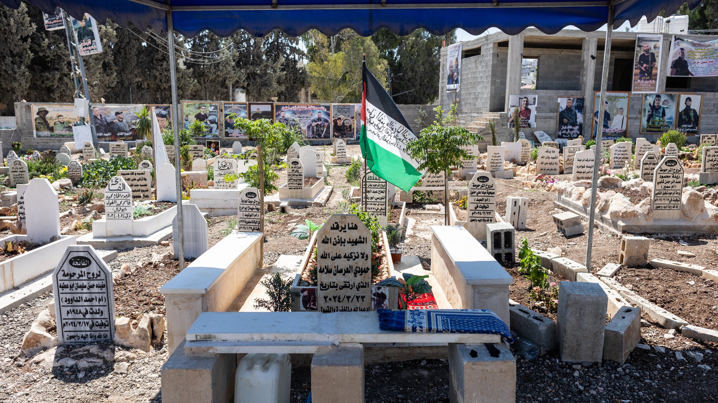 L'immagine mostra un cimitero all'aperto, con diverse tombe disposte in un'area ben curata. Le lapidi sono in varie posizioni e materiali, molte sono contrassegnate con iscrizioni in arabo. Ci sono anche alcune piante verdi e fiori, che aggiungono un tocco di vita al luogo. Al centro, si può vedere una bandiera con i colori della Palestina che sventola, e sullo sfondo ci sono delle fotografie appese, probabilmente in memoria di persone care. Il cimitero è coperto da una tettoia, creando un'area protetta. L'atmosfera è solenne e rispettosa.