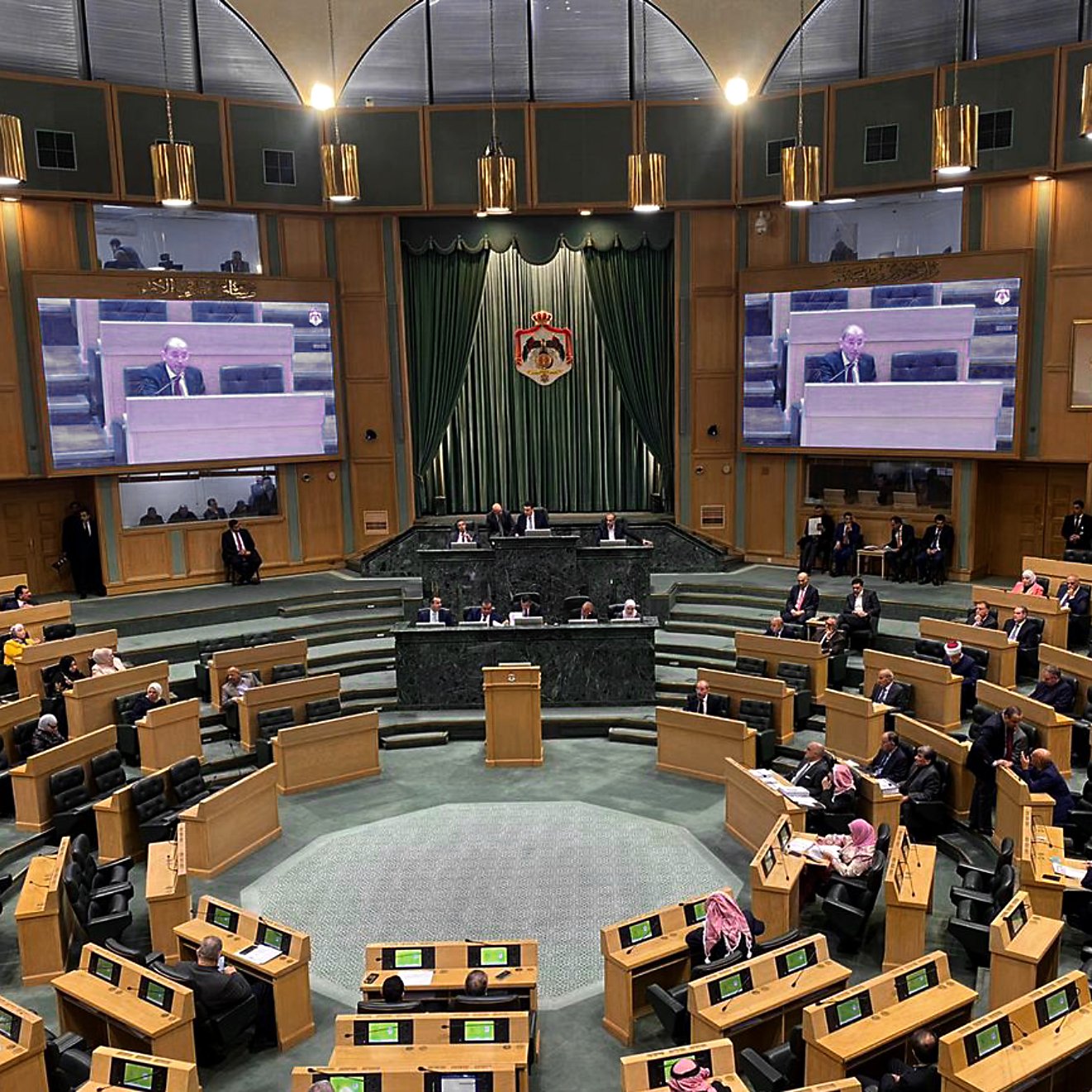 L'image montre l'intérieur d'une grande salle de réunion ou d'assemblée, probablement un parlement ou une assemblée législative. Au centre, une estrade est prévue pour les orateurs, et des écrans sont visibles sur les murs pour diffuser les interventions. On peut voir plusieurs membres assis sur des rangées en pente, organisés en sections. Les couleurs dominantes sont des tons de vert et de bois, créant une ambiance formelle. Des personnes sont également présentes debout, écoutant attentivement.