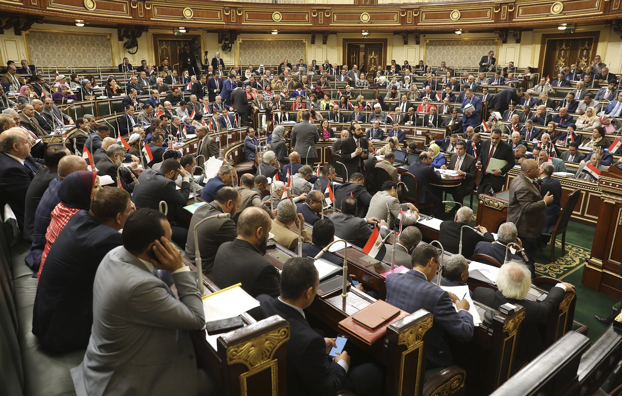 L'image montre une assemblée ou un parlement en pleine session. De nombreux représentants sont assis dans des rangées, certains discutent tandis que d'autres prennent des notes ou consultent des documents. On peut voir des drapeaux égyptiens sur certaines tables, ce qui suggère que cette image provient de l'Assemblée nationale d'Égypte. L'environnement est formel, avec une riche décoration et une disposition typique d'une salle de réunion politique.