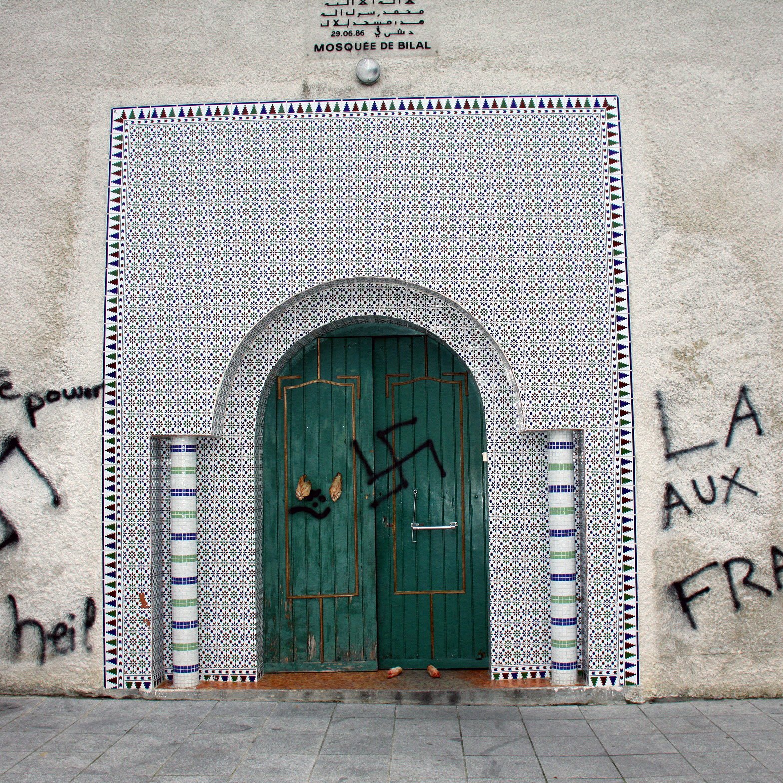 L'image montre un mur extérieur d'une mosquée, orné de carreaux colorés. Sur le mur, il y a des graffitis qui incluent un symbole controversé, représentant une swastika, ainsi que des phrases telles que "White power" et "La France aux Français." La porte d'entrée est en bois, avec des détails architecturaux typiques. L'ensemble de la scène évoque un message de haine et de division.