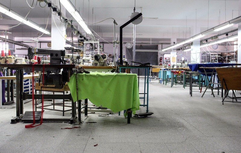 تظهر الصورة مشهدًا داخليًا لورشة خياطة. المساحة تبدو واسعة ومشرقة، حيث توجد آلات خياطة متعددة منتشرة في المكان. بعض الطاولات تحتوي على أقمشة ملونة، في حين أن العديد من المعدات والأدوات موجودة في حالة استخدام أو جاهزية. الأرضية مغطاة ببعض القصاصات من القماش، مما يشير إلى نشاط العمل. الإضاءة تأتي من الأنوار السقفية، وهناك ساعة على الحائط تدل على الوقت. يبدو أن الورشة تحتاج إلى بعض الترتيب.