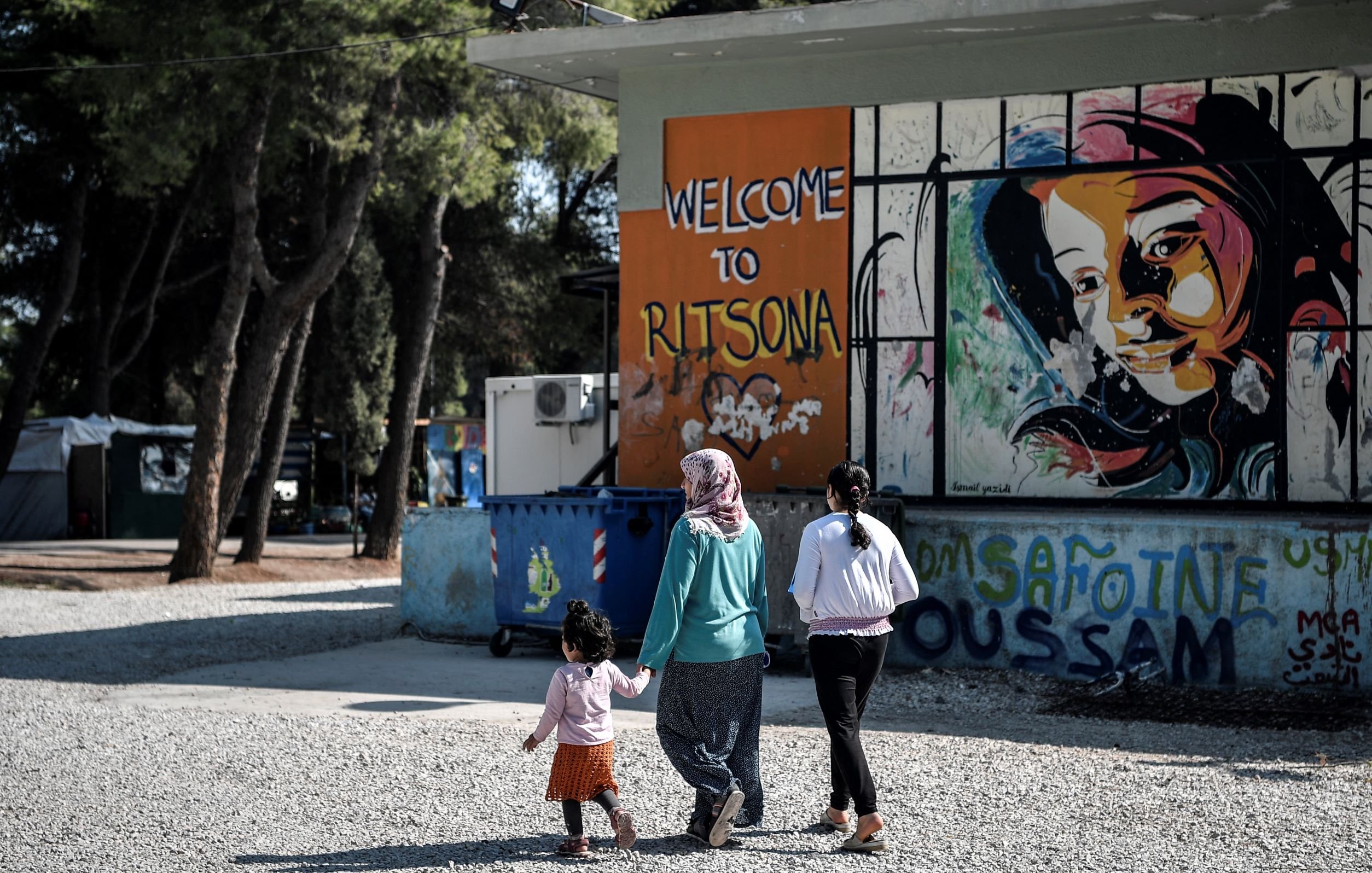 L'image montre un groupe de personnes marchant sur un chemin en gravier. À gauche, on peut voir un conteneur à ordures et à droite, un mur peint avec des graffitis colorés, dont le mot "Welcome to Ritsona" et le portrait d'une fille. Les personnes présentes semblent être une mère accompagnée de deux enfants, l'un d'eux tenant la main de la femme. L'environnement végétal suggère un cadre tranquille, malgré la situation.
