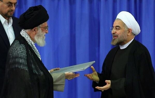 L'image montre deux hommes en tenue traditionnelle, se tenant debout l'un en face de l'autre. L'un d'eux porte un turban noir et une cape sombre, tandis que l'autre est vêtu d'une robe noire avec un col ouvert. Ils semblent être en train d'échanger un document ou un dossier, avec des expressions de concentration sur leurs visages. L'arrière-plan est de couleur bleue, ce qui pourrait suggérer un cadre officiel ou un événement marquant.