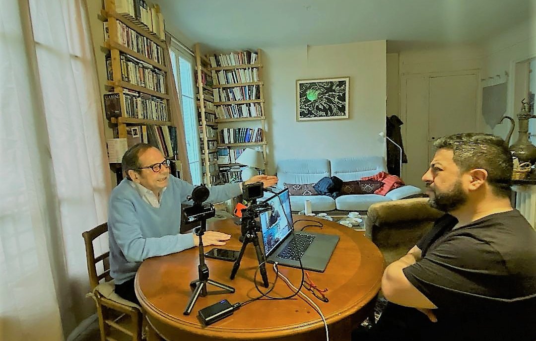 The image depicts two men engaged in a conversation at a round wooden dining table. One man, wearing a light blue sweater and glasses, appears to be animatedly speaking and gesturing, while the other man, dressed in a black shirt, is listening attentively. On the table, there are microphones and a laptop, suggesting they might be recording a podcast or conducting an interview. In the background, a cozy living space is visible with shelves full of books and a sofa where another person seems to be resting. The overall atmosphere feels informal and collaborative.
