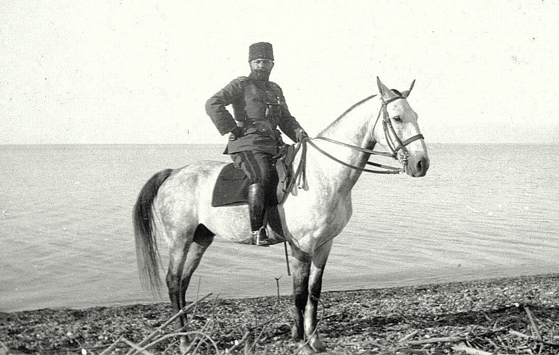 L'image montre un homme en uniforme militaire monté sur un cheval blanc. Il se tient debout sur la plage, près d'une étendue d'eau. L'homme porte un couvre-chef distinctif, et son uniforme semble traditionnel. Le paysage est simple, avec des galets au premier plan et un horizon calme en arrière-plan. L'atmosphère donne une impression de sérénité et de majesté.