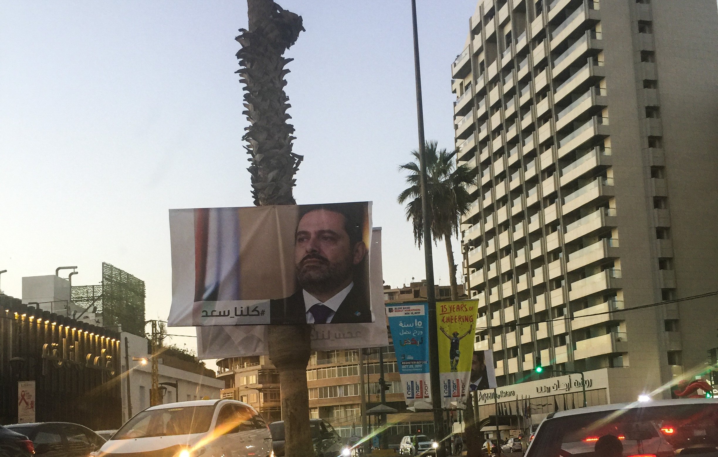 L'image montre une vue urbaine avec un grand panneau publicitaire affichant le visage d'un homme en costume, probablement un homme politique, et des arbres de palmiers sur le côté. En arrière-plan, on peut voir un immeuble de grande hauteur avec des enseignes, et d'autres panneaux d'affichage autour. La lumière du soir donne une ambiance douce à la scène. Les voitures sont visibles sur la route, suggérant une circulation urbaine.