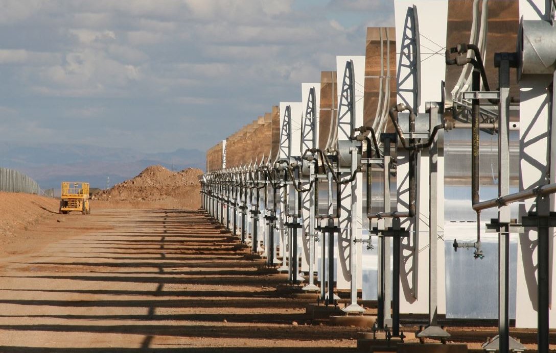 L'image montre une série de panneaux solaires en concentrateurs, alignés sur un terrain désertique. Ces structures lumineuses captent la lumière du soleil pour générer de l'énergie. En arrière-plan, on peut voir un ciel partiellement nuageux avec des montagnes lointaines. L'environnement semble aride, avec des collines de terre et un véhicule de construction visible à distance. Les ombres des panneaux s'étendent sur le sol, soulignant leur orientation vers le soleil.