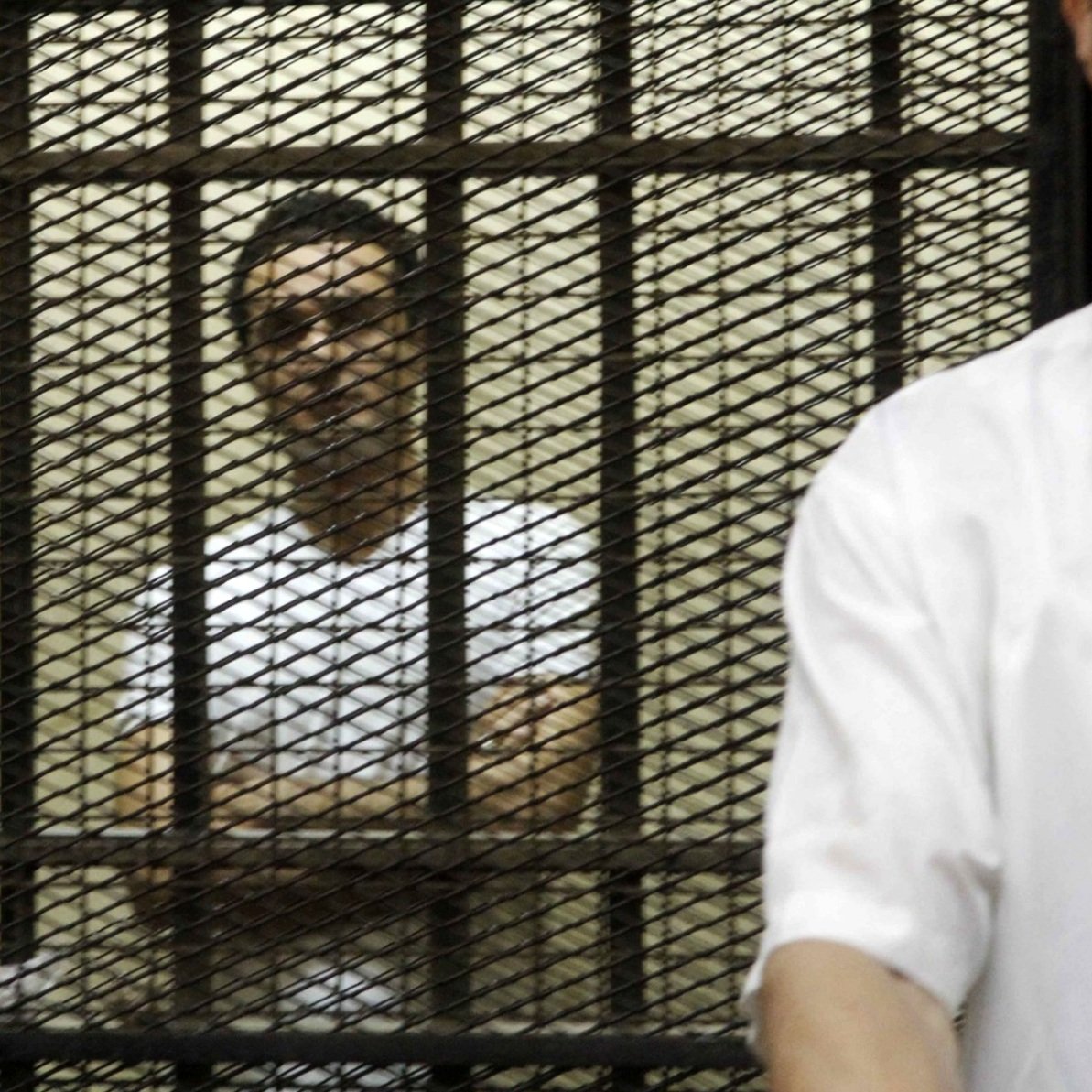 Deux hommes se tiennent derrière des barreaux, l'un visible et l'autre en arrière-plan.