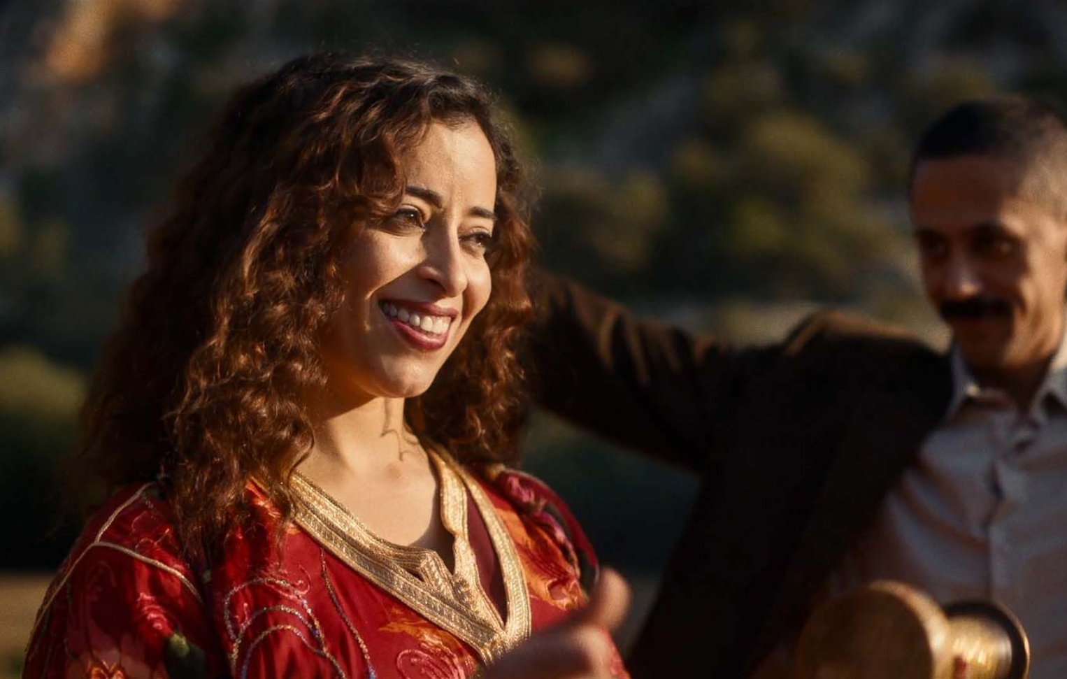 L'image montre une femme souriante avec des cheveux bouclés, portant une robe rouge ornée de motifs. Elle semble joyeuse et est entourée d'un environnement naturel. Un homme en arrière-plan, vêtu d'un costume, semble également profiter du moment. L'atmosphère générale est festive et conviviale.