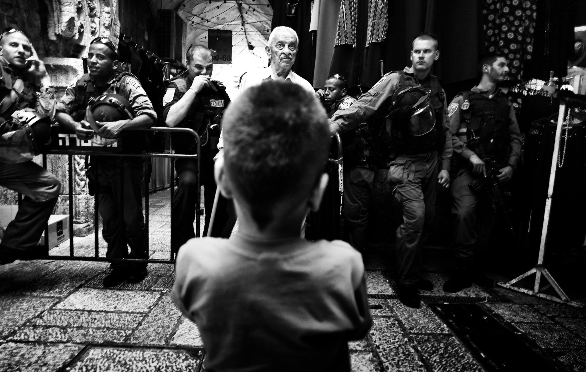 L'image montre un enfant vu de l'arrière, regardant vers un groupe de personnes, dont certaines semblent être des policiers ou des soldats. L'atmosphère est tendue, avec des barrières de sécurité en place et une composition en noir et blanc qui accentue le contraste entre les figures. L'enfant, central dans la scène, semble intrigué ou préoccupé par la situation qui se déroule devant lui. Les autres personnes sont alignées, probablement en position d'intervention.
