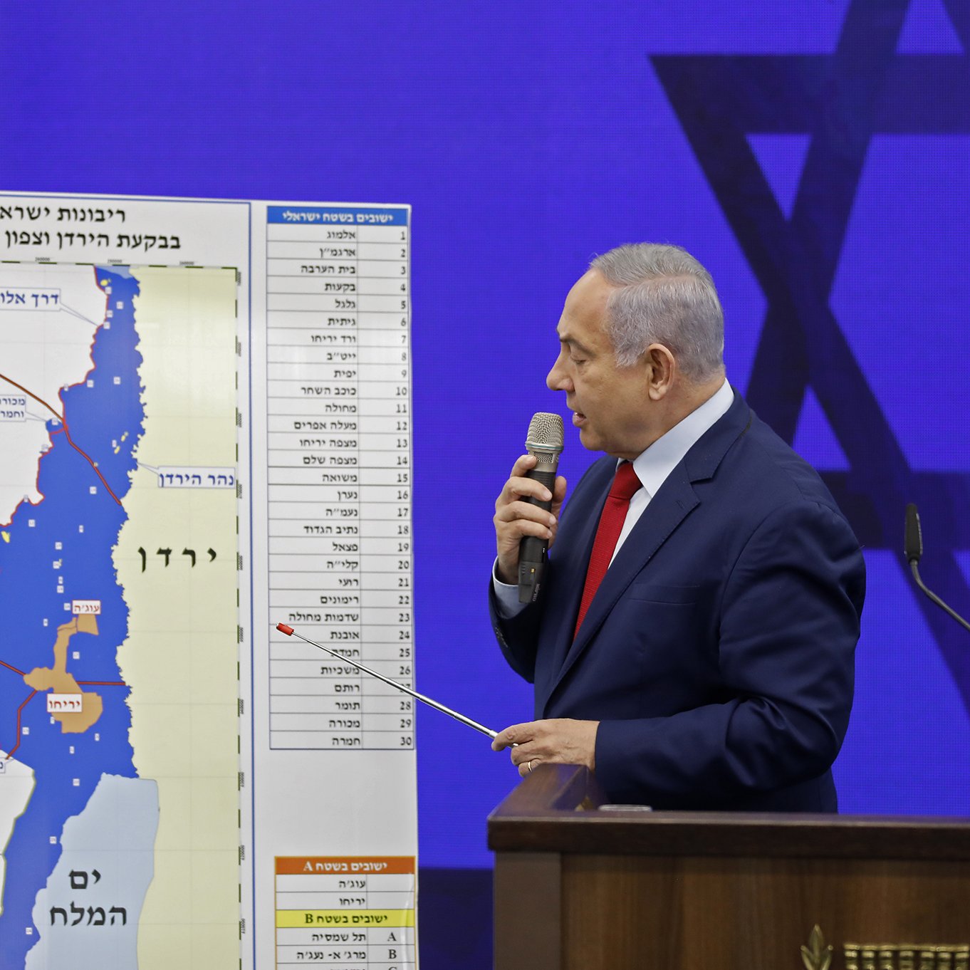 L'image montre un homme debout devant un tableau graphique. Il semble s'adresser à un auditoire, tenant un pointeur dans sa main. Le tableau derrière lui illustre une carte, probablement liée à une question de territoire, avec des annotations en hébreu. On aperçoit également un symbole du judaïsme en arrière-plan. L'homme porte une cravate rouge et un costume sombre.