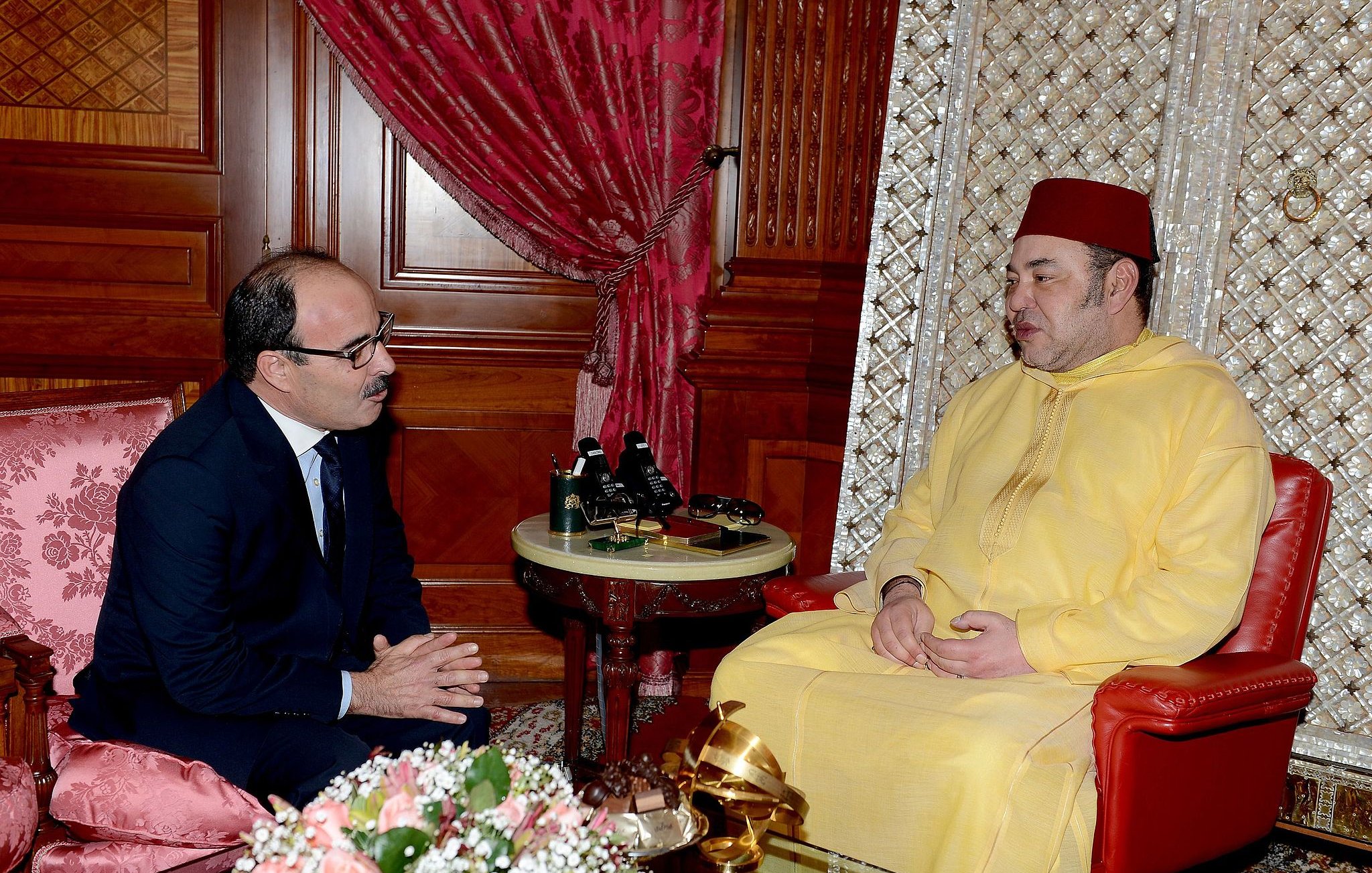 L'image montre deux hommes assis dans une pièce richement décorée. L'un est vêtu d'un habit traditionnel jaune et porte un fez, tandis que l'autre est en costume. Ils sont engagés dans une conversation, avec des expressions sérieuses. En arrière-plan, on peut voir des éléments décoratifs, tels que des rideaux rouges et un arrangement floral sur une table.