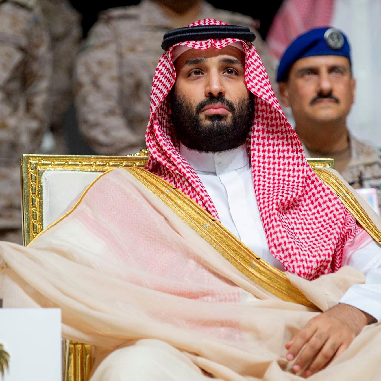 Homme en tenue traditionnelle, assis, regard sérieux, décor militaire en arrière-plan.