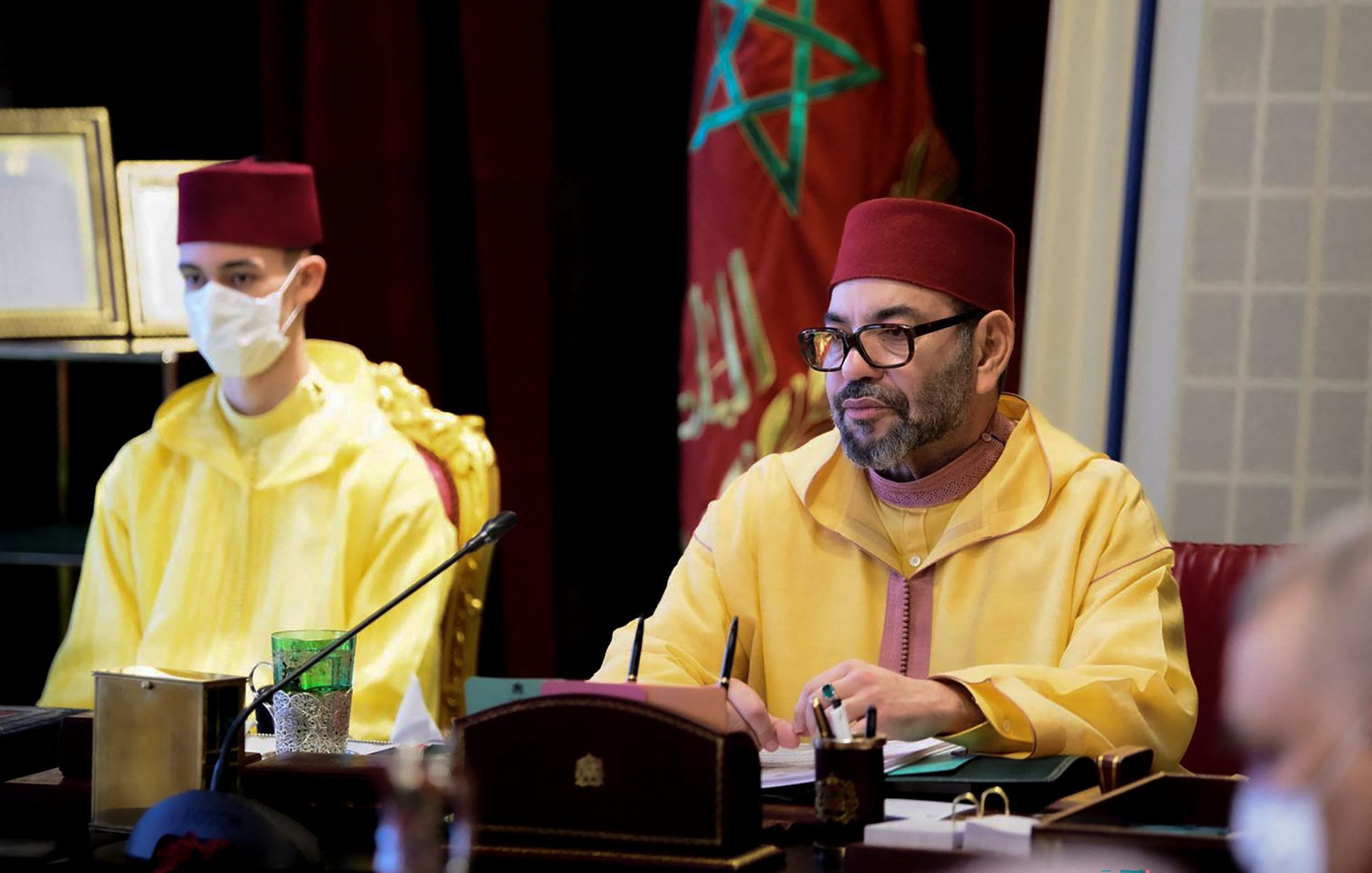 L'immagine mostra un incontro ufficiale in corso. A sinistra, un giovane indossa un abito giallo e una mascherina, mentre a destra c'è un uomo con un berretto rosso e un abbigliamento tradizionale marocchino di colore giallo. L'uomo a destra sembra concentrato, forse mentre prende appunti o partecipa a una discussione. Sullo sfondo si intravedono elementi decorativi di un ambiente formale.