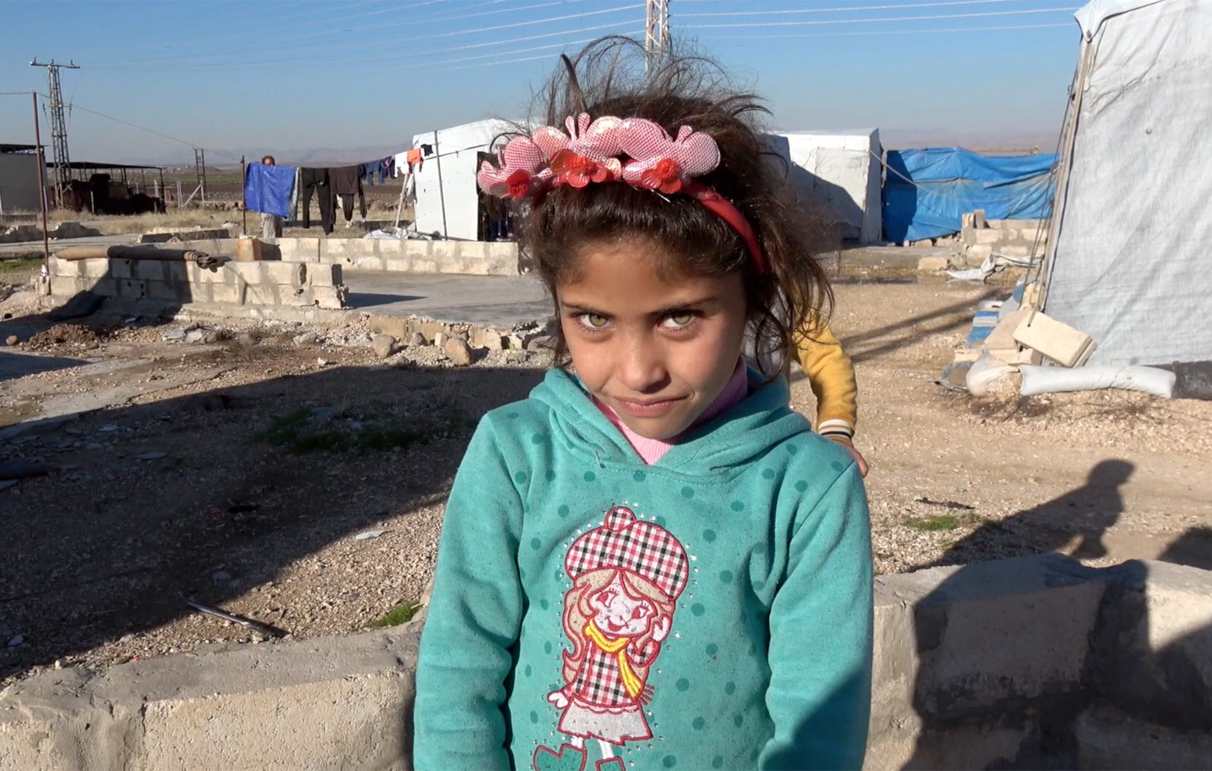 The image depicts a young girl with striking blue eyes and dark hair, wearing a bright turquoise sweatshirt adorned with a character design on the front. She has a decorative pink headband with flowers, which adds a playful touch to her appearance. The background shows a makeshift camp with tents and some structures, indicating a setting that may be temporary or in a challenging environment. The girl is looking directly at the camera with a slight smile, suggesting a sense of resilience and charm amidst her surroundings.